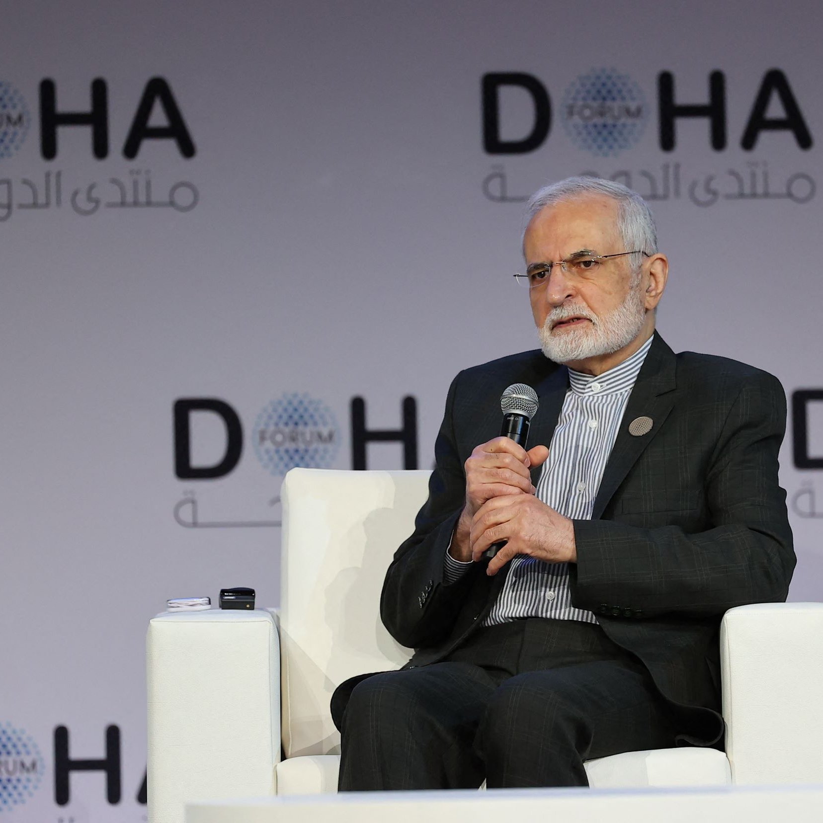 L'image montre un homme assis sur un fauteuil blanc, participant à une conférence ou un forum. Il porte une tenue formelle et semble être en train de s'exprimer ou d'écouter. En arrière-plan, on peut voir un logo avec le mot "DOHA" et d'autres inscriptions en arabe. Des fleurs et une bouteille d'eau sont également visibles sur la scène. L'atmosphère semble professionnelle et tranquille.