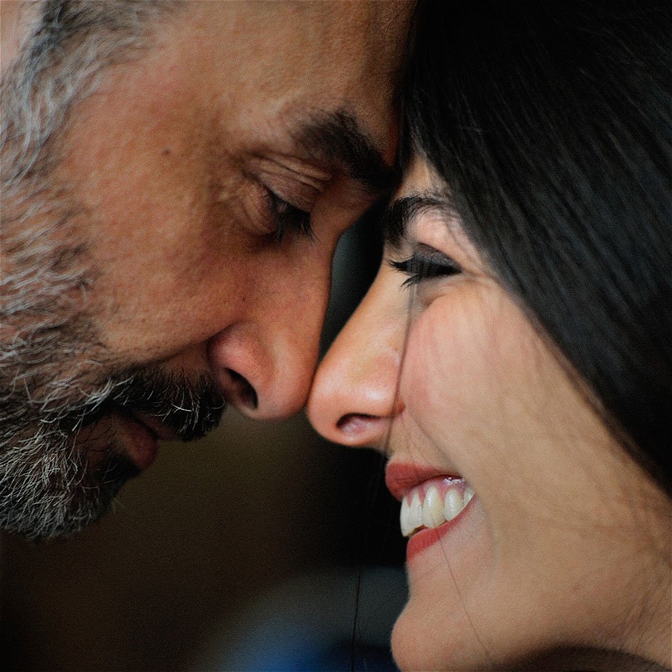 Cette image montre deux personnes, un homme et une femme, qui sont très proches l'un de l'autre. Ils touchent leurs fronts et semblent partager un moment intime. L'homme a une barbe grisonnante, tandis que la femme a des cheveux longs et foncés. Leur expression est douce : l'homme a un air serein, et la femme sourit joyeusement. L'ambiance de l'image évoque l'affection et la complicité entre les deux individus.