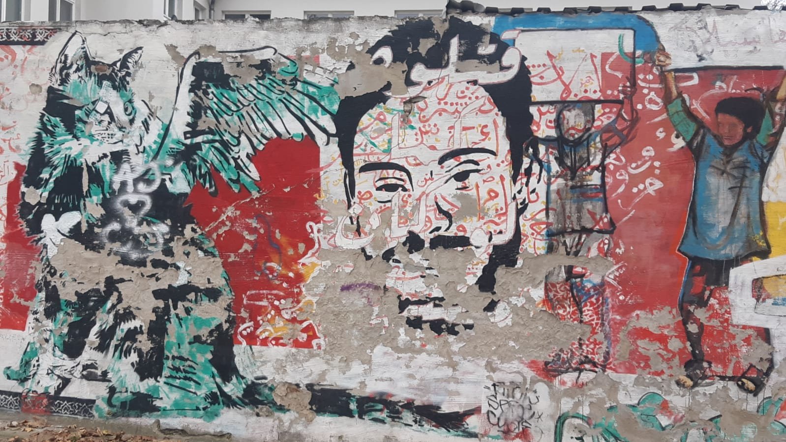 L'image montre une fresque murale riche en couleurs et en détails. À gauche, on peut voir un personnage avec des ailes et un visage expressif, qui semble être une représentation stylisée d'une créature. À droite, se trouve un visage humain qui attire l'attention, entouré de motifs vibrants. En arrière-plan, divers éléments graphiques et des inscriptions en arabe ajoutent une dimension culturelle. L'ensemble donne une impression de dynamisme et de créativité, typique de l'art urbain.