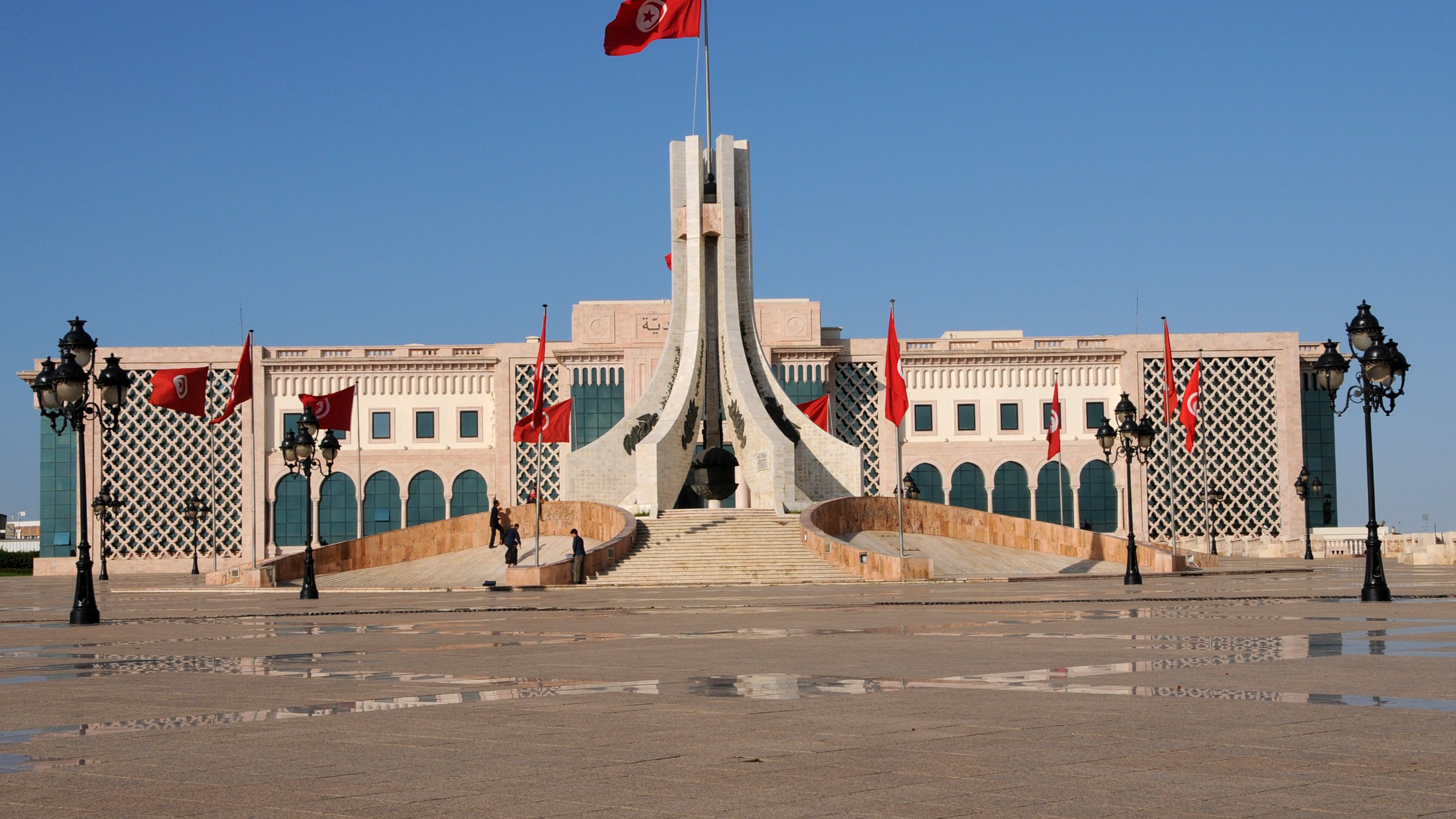 L'image montre un bâtiment imposant avec une architecture distinctive, situé probablement dans un espace public. Au centre, on peut voir un mât portant le drapeau tunisien, avec plusieurs drapeaux rouges autour. L'esplanade devant le bâtiment est pavée, et des lampadaires élégants sont présents sur les côtés. Le ciel est dégagé, ce qui donne une atmosphère lumineuse et claire à la scène. L'architecture semble moderne mais avec des éléments traditionnels.