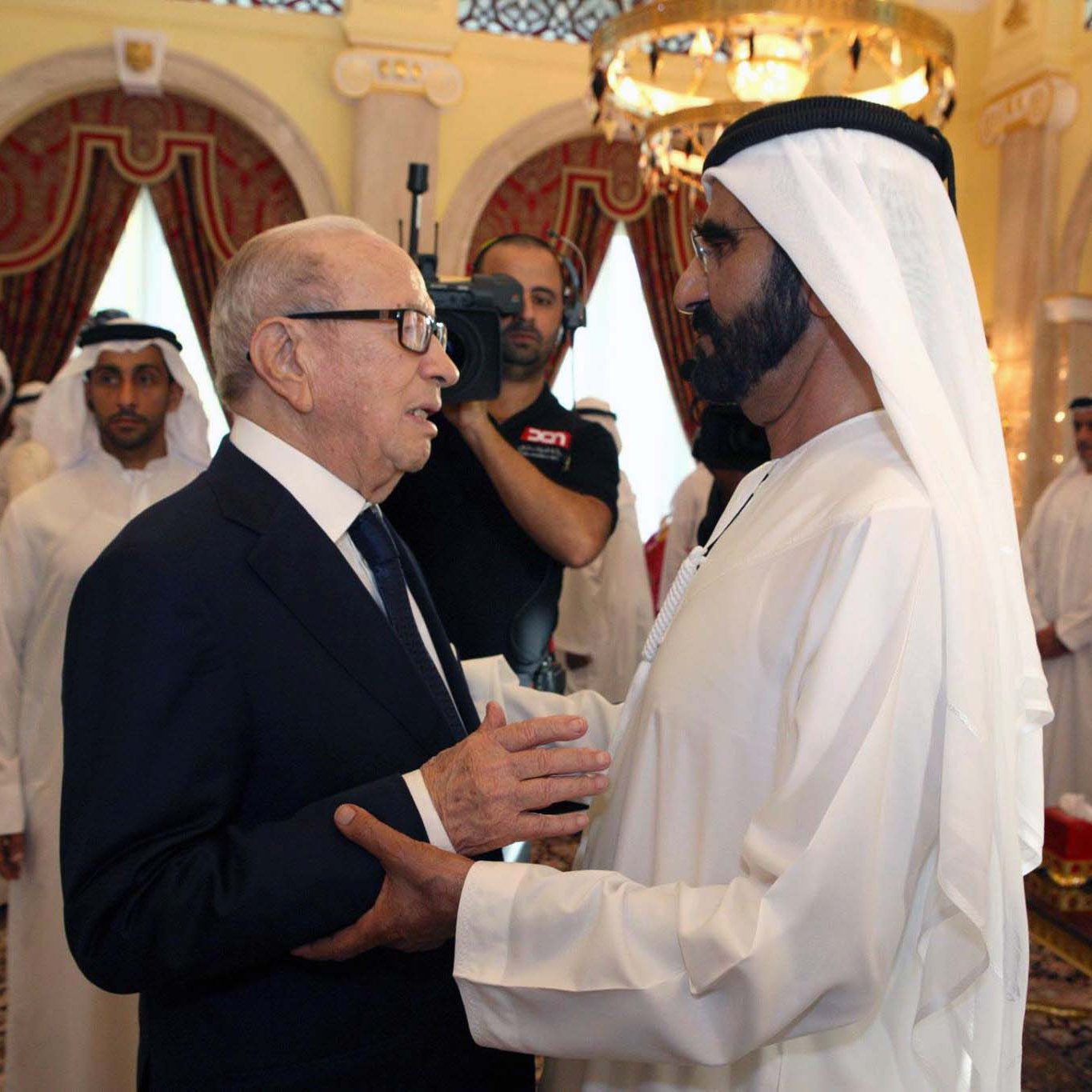 L'image montre deux hommes en discussion, probablement dans un cadre formel et décoré, avec des éléments architecturaux typiques d'un palais. L'un des hommes porte un costume sombre, tandis que l'autre est vêtu d'une robe traditionnelle blanche. En arrière-plan, on aperçoit d'autres personnes, également en tenue traditionnelle, qui semblent observer la conversation. L'ambiance semble conviviale et respectueuse.