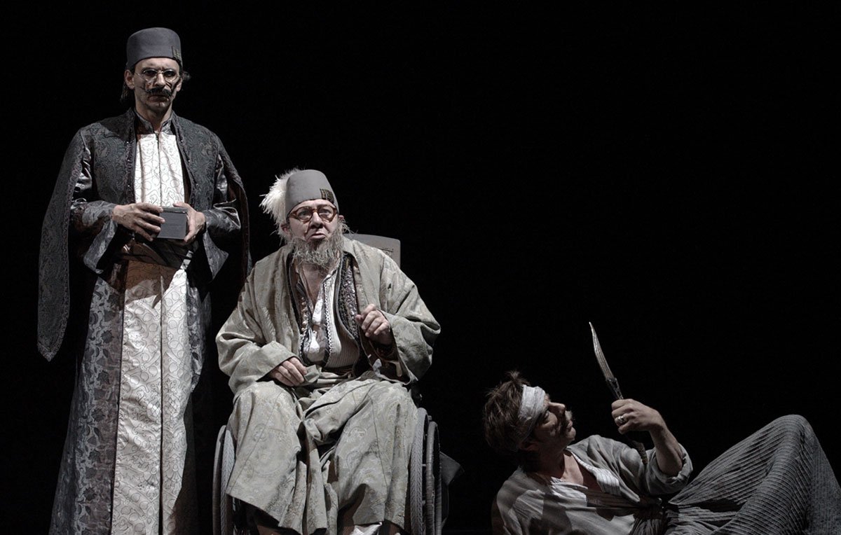 L'image représente une scène de théâtre mettant en avant trois personnages. Deux d'entre eux, habillés de façon traditionnelle et avec des vêtements sombres et ornés, semblent être en position d'autorité ou d'observation. Le troisième personnage est assis dans un fauteuil roulant, avec des vêtements plus clairs et un air pensif ou mélancolique. Il a une longue barbe grise et des lunettes. À côté de lui, un autre personnage, qui paraît être en détresse ou blessé, est au sol, tenant un objet pointu. L'éclairage est sombre, créant une atmosphère dramatique et intense.