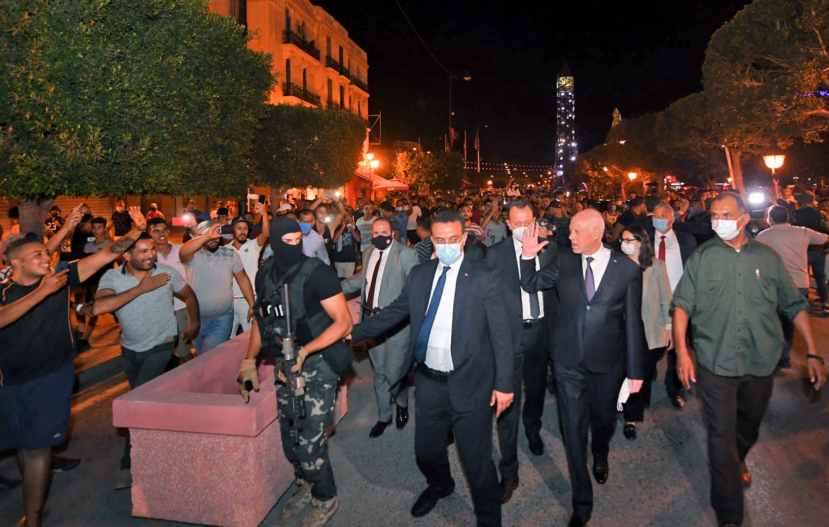 La imagen muestra a un grupo de personas en una calle durante la noche. En el centro, hay un hombre vestido con traje y corbata, escoltado por un guardia de seguridad con una máscara y uniforme. Alrededor, se ven varios manifestantes levantando las manos, algunos parecen estar enérgicos o en protesta. La iluminación de la escena proviene de faroles y luces en el entorno, creando un ambiente animado y posiblemente tenso. Los árboles y edificios en el fondo sugieren que se trata de una zona urbana.