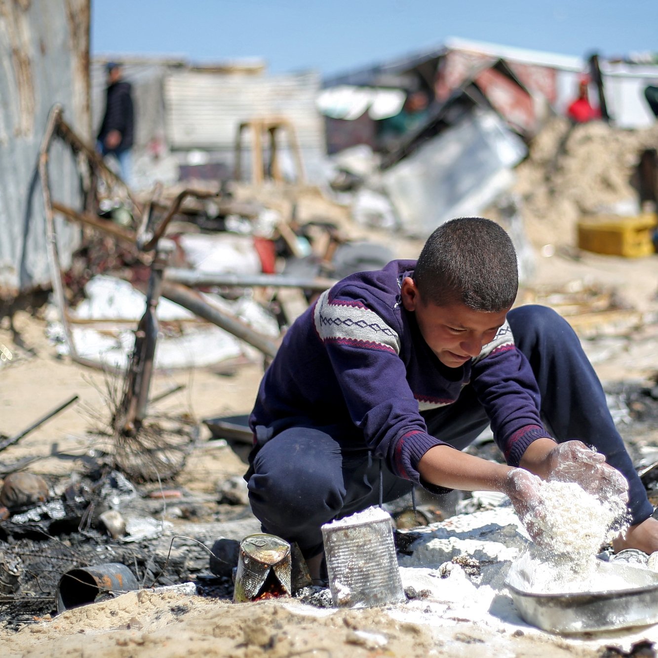پسرکی در میان خرابه‌ها نشسته و مشغول بازی با مواد سفید رنگ است. فضای اطراف بسیار بی‌نظم و ویران است.