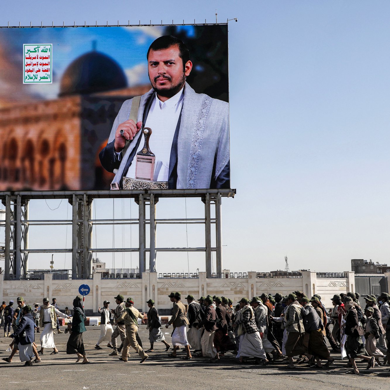 L'image montre un grand panneau d'affichage avec un portrait d'un homme en tenue traditionnelle, probablement un leader ou une figure politique, devant un paysage architectural. En dessous, un groupe de personnes marche en procession, portant des drapeaux et des affiches. L'environnement semble urbain, suggérant une manifestation ou un événement public. Les écriteaux sur le panneau en arabe ajoutent au contexte culturel et politique de l'image.