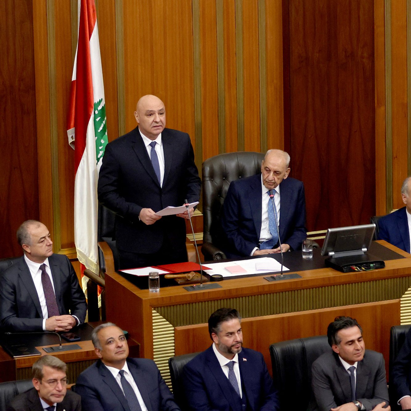 این تصویر یک جلسه رسمی در مجلس لبنان را نشان می‌دهد. در مرکز تصویر، یک فرد در حال سخنرانی است و تعدادی دیگر از اعضای مجلس در اطراف او نشسته‌اند. رومیزی مجلس با مستنداتی پوشیده شده و یک پرچم لبنان نیز در پس‌زمینه قابل مشاهده است. به نظر می‌رسد که جو جلسه جدی و رسمی است.