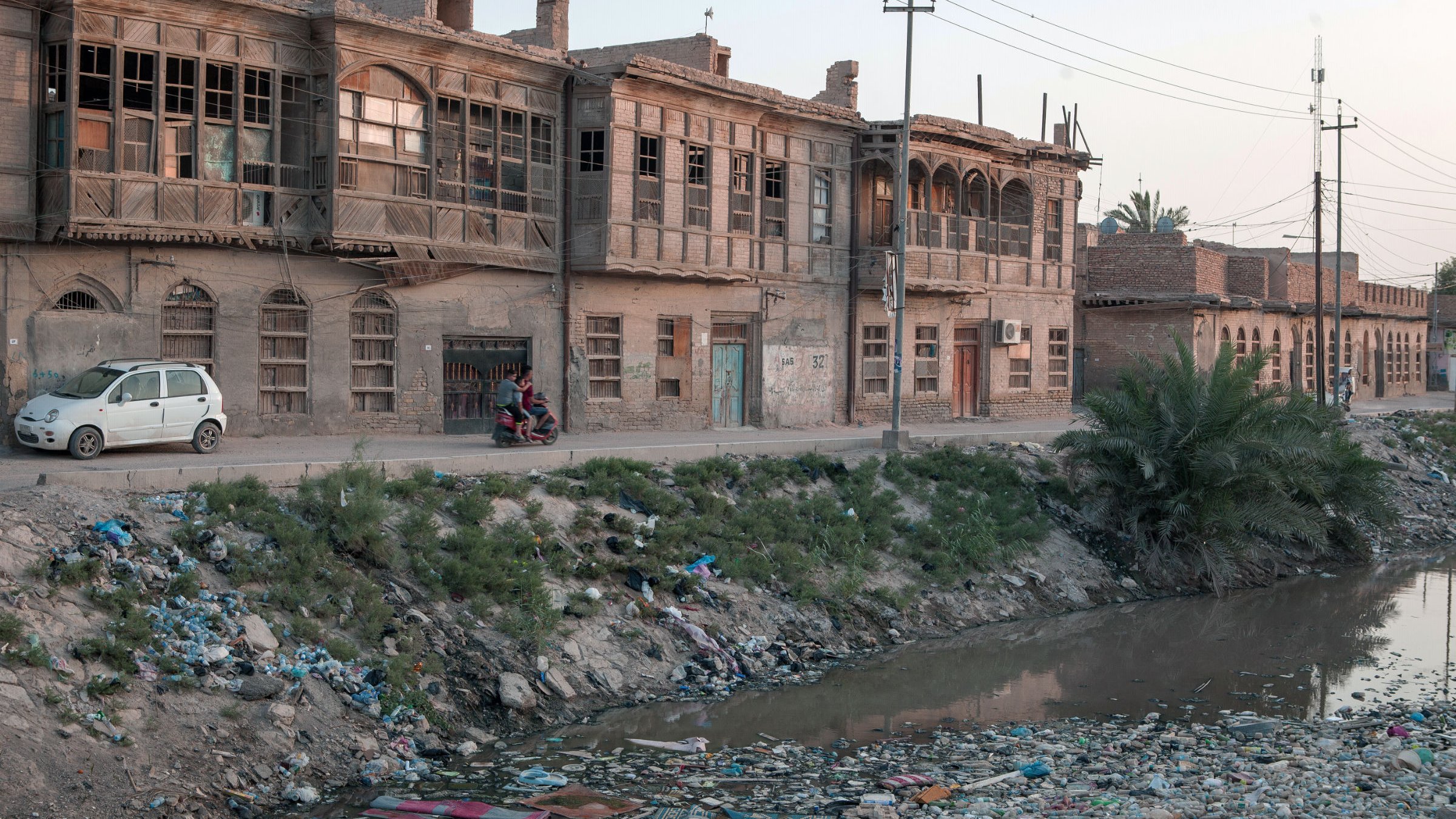 L'image montre une rue bordée de vieilles maisons en briques, caractérisées par des fenêtres en bois. À gauche, on aperçoit une voiture garée. Sur le côté droit, un groupe de personnes semble discuter. Au premier plan, un bras de rivière est visible, rempli de déchets et de débris, entouré de végétation. L'éclairage doux de la fin de journée crée une atmosphère nostalgique dans ce paysage urbain en dégradation.