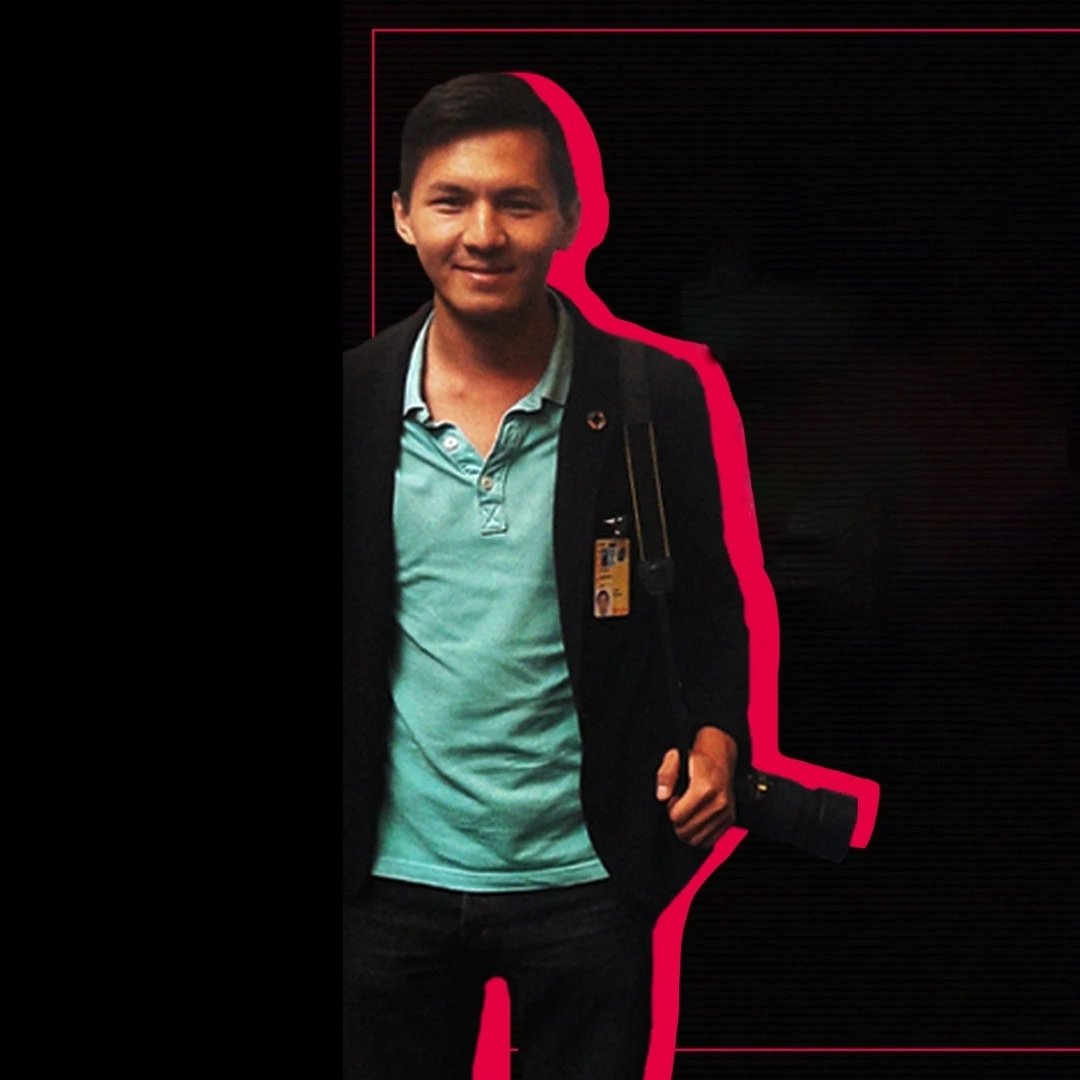 L'image montre un homme souriant, portant une chemise turquoise sous un veston noir. Il a une carte d'identification accrochée à son cou et tient un appareil photo à la main. Le fond est principalement noir, avec un contour rose qui souligne la silhouette de l'homme. En haut à droite, le logo "RSF" est visible.