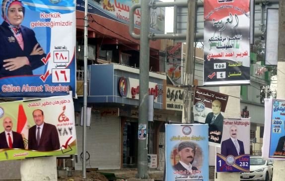 L'image montre une rue animée bordée de panneaux électoraux. On peut y voir plusieurs affiches de campagne, chacune présentant différents candidats avec leurs noms et numéros associés, ainsi que des logos de partis politiques. Les couleurs des affiches varient, attirant l'attention des passants. L'environnement urbain semble typique d'une zone commerçante, avec des boutiques et des enseignes visibles en arrière-plan.