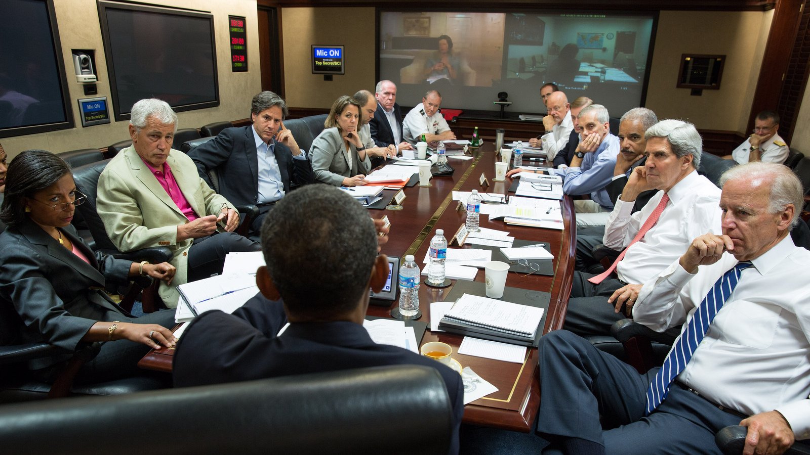 L'image montre une réunion importante autour d'une grande table. Plusieurs personnes, des hommes et des femmes, sont assis, engagés dans une discussion sérieuse. À l'arrière-plan, on peut voir un écran où se tient une vidéoconférence. Les participants semblent être des responsables et des conseillers, probablement dans un contexte gouvernemental ou militaire. L'ambiance générale est concentrée, avec des documents étalés sur la table et des bouteilles d'eau à disposition.