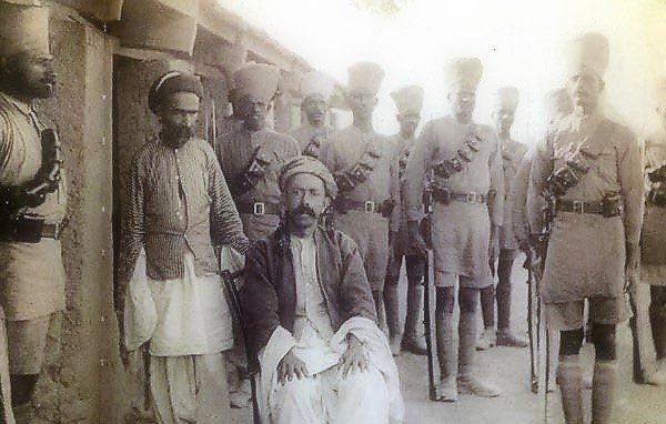 الصورة تظهر مجموعة من الرجال يرتدون زيًا عسكريًا تقليديًا، يقفون في صفوف. في المقدمة، يجلس رجل على كرسي، يبدو أنه شخصية ذات مكانة أو أهمية. يرتدي الرجل ملابس تقليدية مختلفة عن الآخرين، مما يدل على اختلاف اجتماعي أو ثقافي. الخلفية تبدو بسيطة، وتضيف إلى الطابع التاريخي للصورة. الأجواء تتسم بالجدية، ويبدو أن الصورة قد التُقطت في فترة زمنية سابقة.