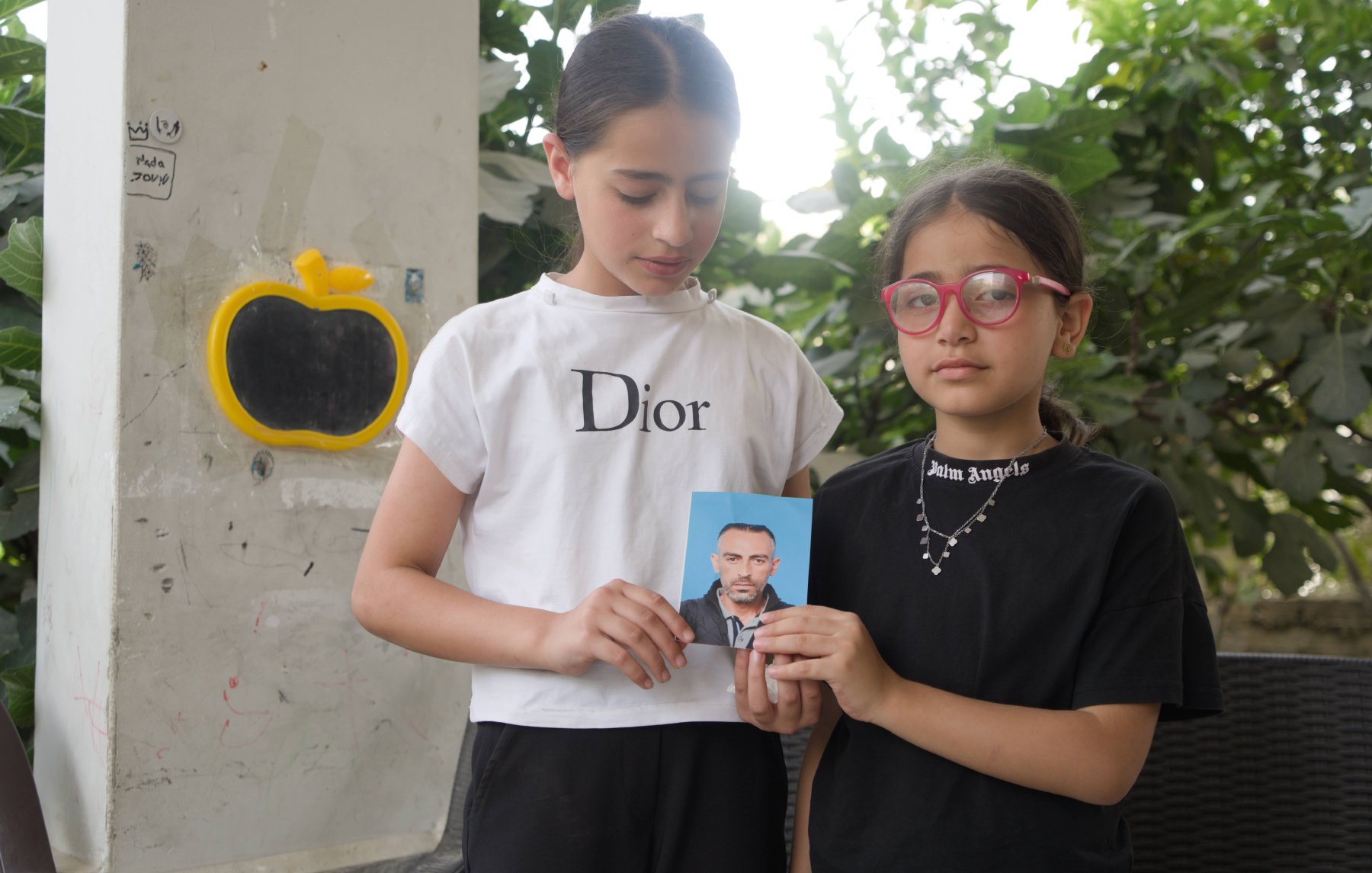 Sur l'image, on voit deux jeunes filles debout l'une à côté de l'autre. La fille de gauche porte un t-shirt blanc avec le logo "Dior" et tient en main une photo d'un homme. La fille de droite est vêtue d'un t-shirt noir avec des lunettes roses. Elles sont entourées par un arrière-plan verdoyant, avec des feuilles visibles autour d'elles.
