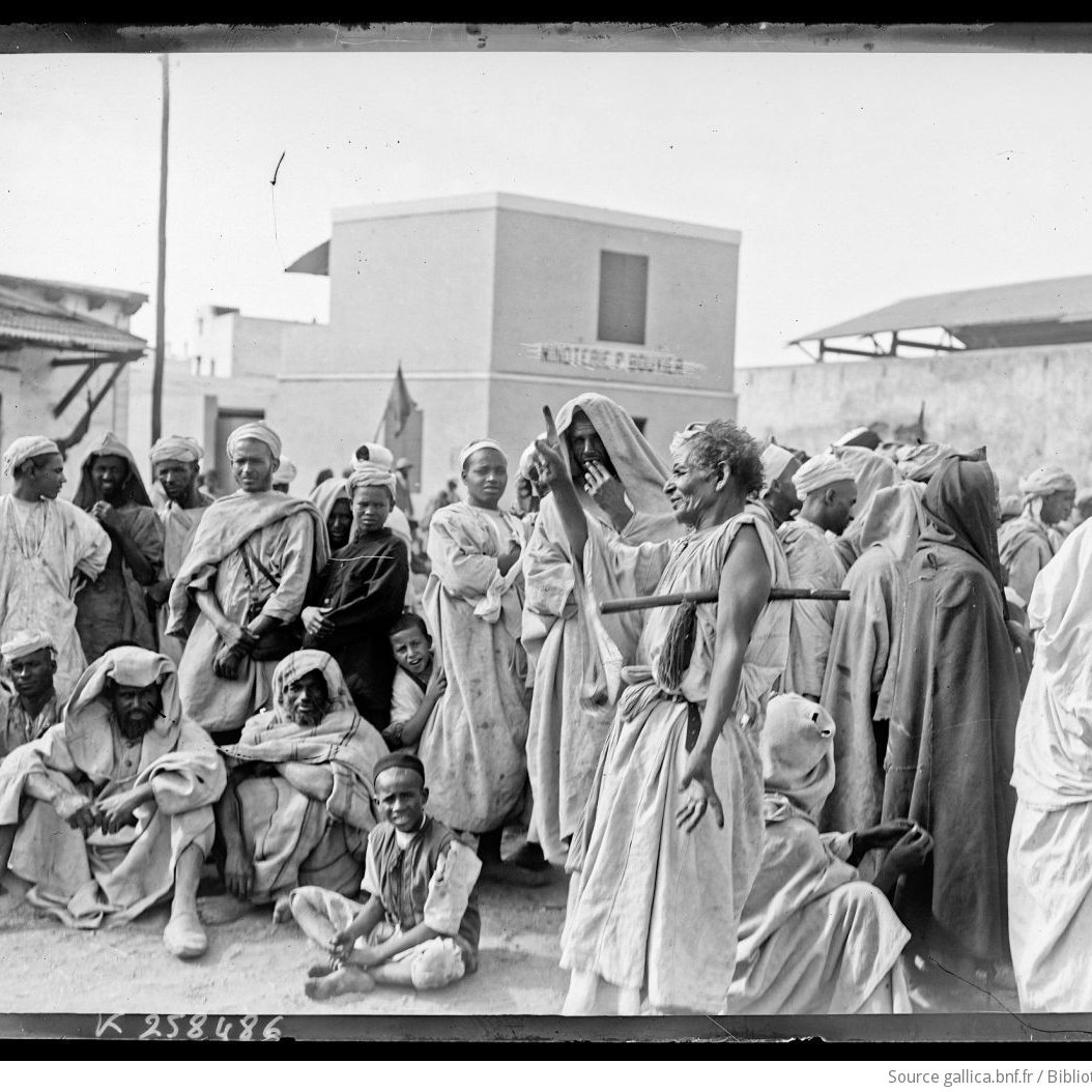 تظهر الصورة مشهداً تاريخياً يضم مجموعة من الأشخاص يرتدون ملابس تقليدية في بيئة حضرية. يتواجد الرجال والنساء بشكل مختلط، بعضهم يجلس وآخرون واقفون، حيث يبدو أنهم يعيشون لحظة تتسم بالنقاش أو الاحتجاج. الخلفية تضم مباني بسيطة، مما يشير إلى بيئة قد تكون في منطقة شمال إفريقيا أو الشرق الأوسط. تعكس الملابس والتعبيرات الثقافية تفاصيل الحياة الاجتماعية في تلك الفترة.