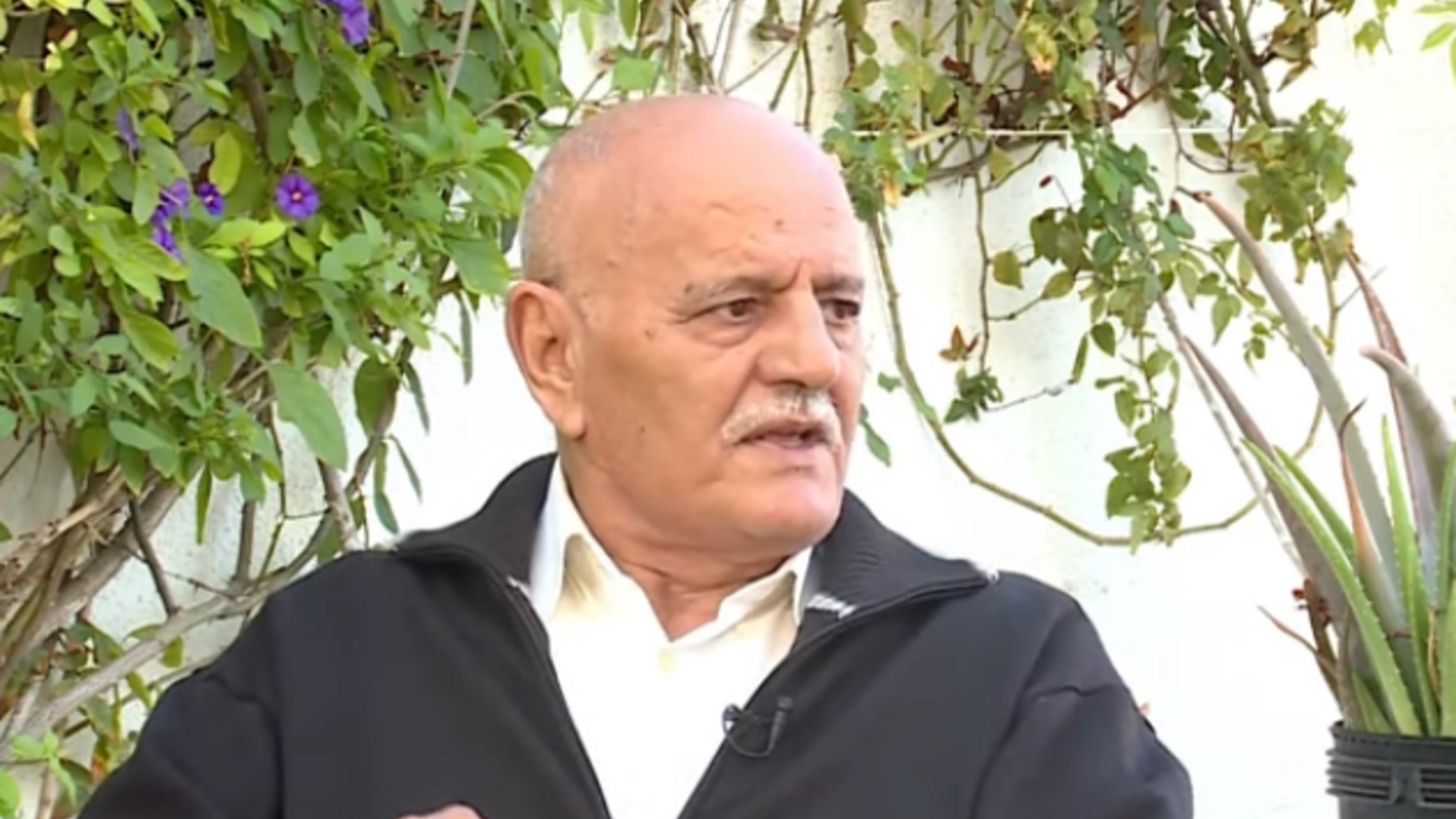 تظهر الصورة شخصًا في منتصف العمر يجلس في مكان خارجي، محاطًا بالنباتات الخضراء. يرتدي سترة داكنة ولديه لحية وشعر رمادي. يبدو وكأنه يتحدث أو يعبّر عن رأيه في موضوع ما، حيث يرفع إصبعه للتأكيد على نقطة سابقة. الخلفية تحتوي على نباتات وأزهار، مما يضفي جوًا طبيعيًا على المشهد.