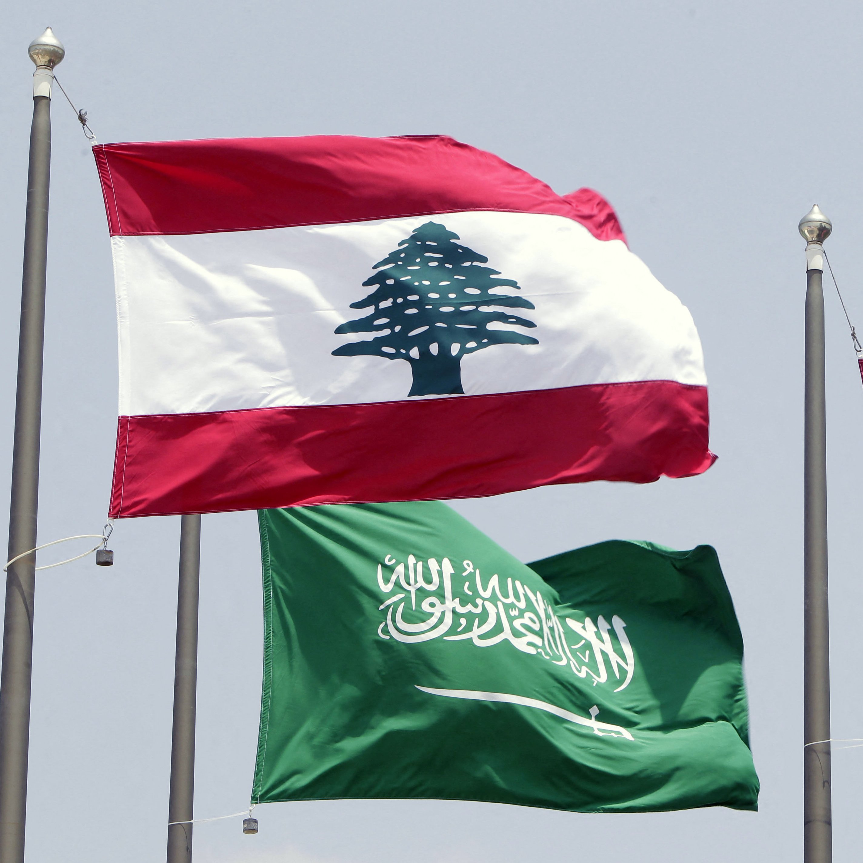 L'image montre deux drapeaux flottant côte à côte. Le drapeau du Liban est en haut, avec une bande rouge en haut et en bas, et une grande illustration d'un cèdre vert au centre. En dessous, le drapeau de l'Arabie Saoudite est vert, portant une inscription en arabe et une épée blanche. Les drapeaux sont hissés sur des mats, probablement lors d'un événement officiel ou d'une rencontre diplomatique.