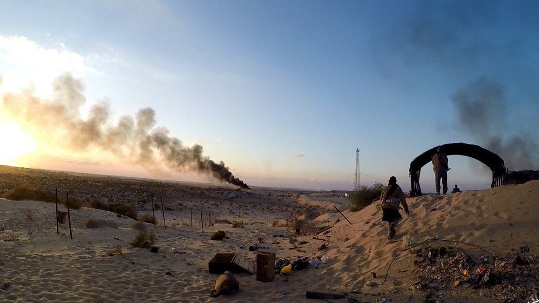 L'image montre un paysage désertique avec une ambiance dramatique. À l'arrière-plan, on peut voir des colonnes de fumée noire s'élevant dans le ciel, suggérant qu'il y a un incendie ou une explosion. Au premier plan, des silhouettes de personnes évoluent dans le sable, probablement en train d'explorer ou de surveiller la zone. La lumière dorée du coucher de soleil crée un contraste frappant avec l'obscurité de la fumée, ajoutant à l'atmosphère générale de tension et de désolation. Le décor semble abandonné, avec quelques débris éparpillés sur le sol.