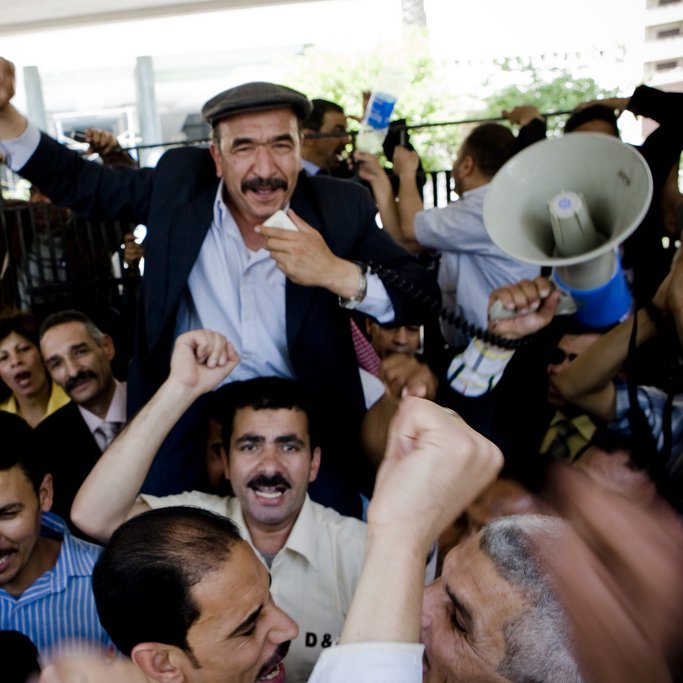 L'image montre une grande foule de personnes rassemblées, manifestant avec enthousiasme. Au centre, un homme souriant est porté sur les épaules d'un autre, levant le poing en signe de victoire. Les autres participants expriment également leur excitation, certains levant les bras et criant. La scène semble vibrante et dynamique, indiquant un moment de célébration ou de rassemblement collectif. Des panneaux et un mégaphone sont également visibles, suggérant une forme de discours ou de protestation.