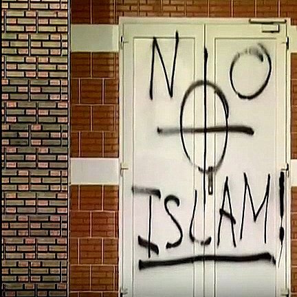 در این تصویر، درب یک ساختمان با رنگ سیاه نوشته شده است. در وسط درب علامت دایره‌ای با خطی عمودی و افقی وجود دارد که به همراه نوشته "NO ISLAM!" که به معنای "اسلام نه!" است، روی درب دیده می‌شود. این نوشته به روشنی یک پیام اعتراضی یا نفرت‌آمیز را منتقل می‌کند. زمینه دیوار نیز آجری و با رنگ‌های متفاوت است.