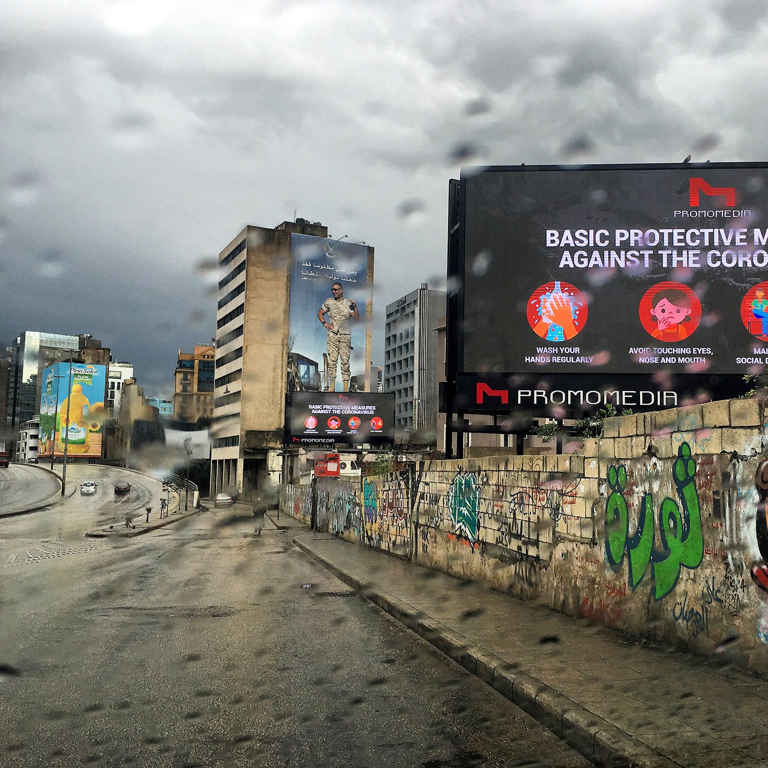 L'image montre une scène urbaine sous la pluie avec des bâtiments modernes à l'arrière-plan. On peut voir un panneau d'affichage numérique qui présente des mesures de protection contre le coronavirus, avec des icônes illustratives. Le mur à gauche est couvert de graffitis colorés, ajoutant une touche artistique à l'environnement. L'atmosphère est plutôt sombre en raison du temps nuageux et pluvieux.