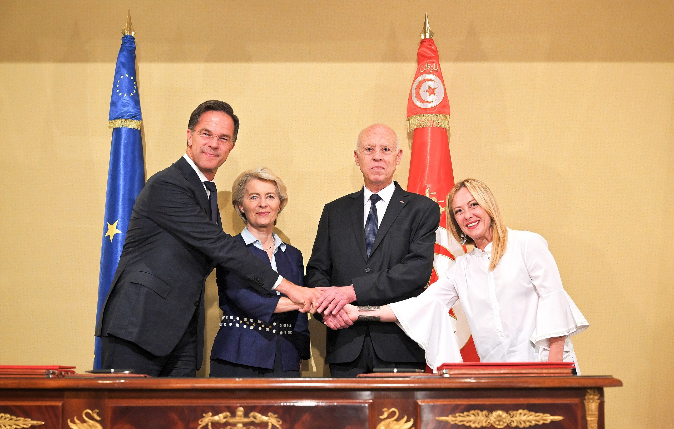 L'image montre quatre personnes se serrant la main autour d'une table. À gauche, un homme en costume sombre, probablement un représentant d'un pays européen. À sa droite, une femme avec des cheveux blonds peignés, semblant être une dirigeante politique. Au centre, un homme en costume sombre, certainement le président d'un pays, et à sa droite, une femme vêtue de blanc, probablement une autre dirigeante. En arrière-plan, on peut voir des drapeaux de l'Union européenne et de la Tunisie, ce qui suggère une rencontre diplomatique.