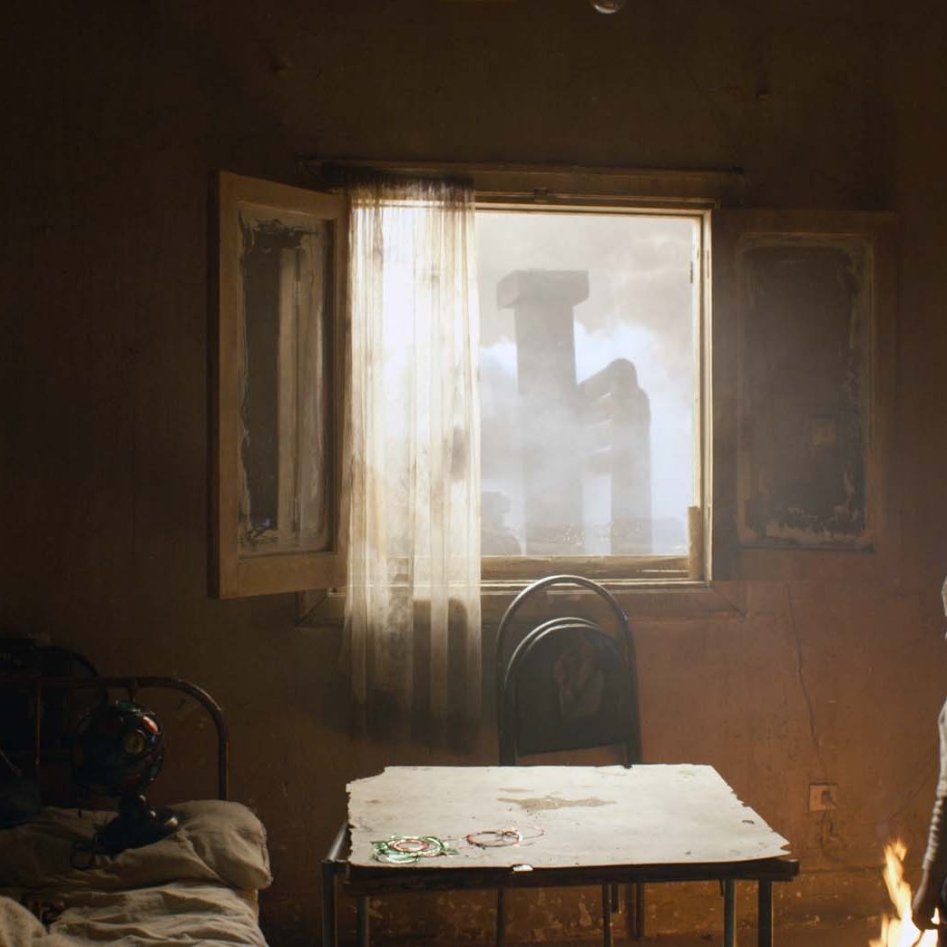 L'image montre une pièce sombre et légèrement en désordre, avec un enfant tourné vers une fenêtre ouverte. La lumière pénètre par cette fenêtre, révélant une scène extérieure brumeuse. À l'intérieur, on voit un lit, une table en bois et un vieux téléviseur. Des décorations festives, comme des ballons et des guirlandes, ajoutent une touche de couleur, bien que l'atmosphère générale semble mélancolique.