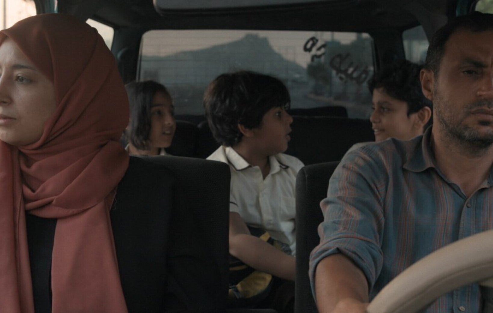 L'image montre un groupe de personnes dans une voiture. À l'avant, un homme au visage sérieux conduit, tandis qu'une femme portant un hijab est assise à côté de lui, semblant pensive. À l'arrière, deux enfants et un adolescent se parlent, créant une atmosphère de dynamisme et d'interaction. La lumière semble douce, suggérant une scène de fin de journée. L'ambiance générale évoque une sorte de voyage ou de déplacement, avec un mélange d'émotions au sein de la voiture.