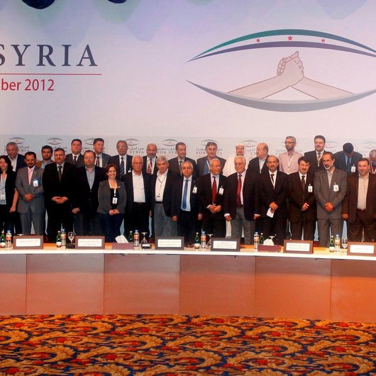 L'image montre un groupe de personnes réunies lors d'un événement formel, probablement une conférence. Elles sont alignées en plusieurs rangées, toutes en costume, et semblent participer à un rassemblement lié à la Syrie, comme indiqué par le texte en arrière-plan. Le décor est coloré, avec des drapeaux et des motifs qui évoquent l'événement. Les participants adoptent des expressions sérieuses, ce qui suggère l'importance de l'occasion.