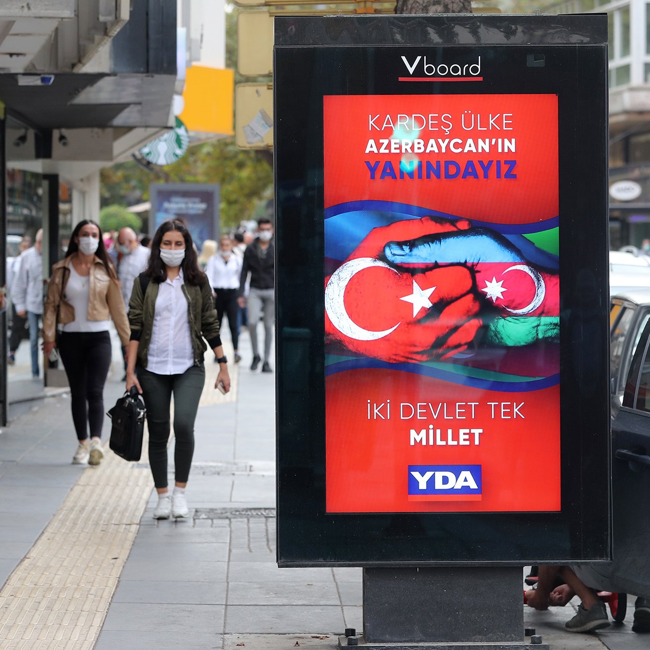 L'image montre une scène de rue animée où des personnes marchent sur un trottoir. À gauche, deux femmes avancent, l'une portant un hijab gris et l'autre en tenue décontractée. À droite, un panneau d'affichage numérique affiche un message en turc, avec des couleurs vives et des symboles représentant les drapeaux de la Turquie et de l'Azerbaïdjan. Le texte semble faire référence à un soutien entre les deux pays. Plusieurs passants peuvent être aperçus en arrière-plan, portant tous des masques de protection.