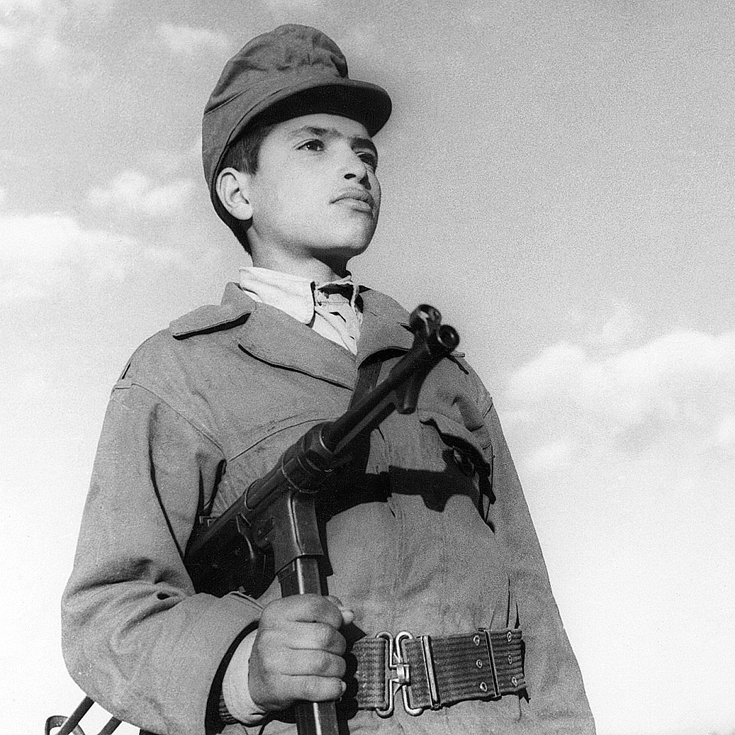 L'image montre un jeune garçon en uniforme militaire, tenant une arme à feu. Son regard est sérieux et déterminé, tandis qu'il se tient droit, face à l'objectif. Le ciel en arrière-plan est clair, avec quelques nuages. L'uniforme et l'arme peuvent indiquer qu'il fait partie d'un contexte militaire ou historique spécifique. L'ambiance de la photo évoque un sentiment de courage et de bravoure.