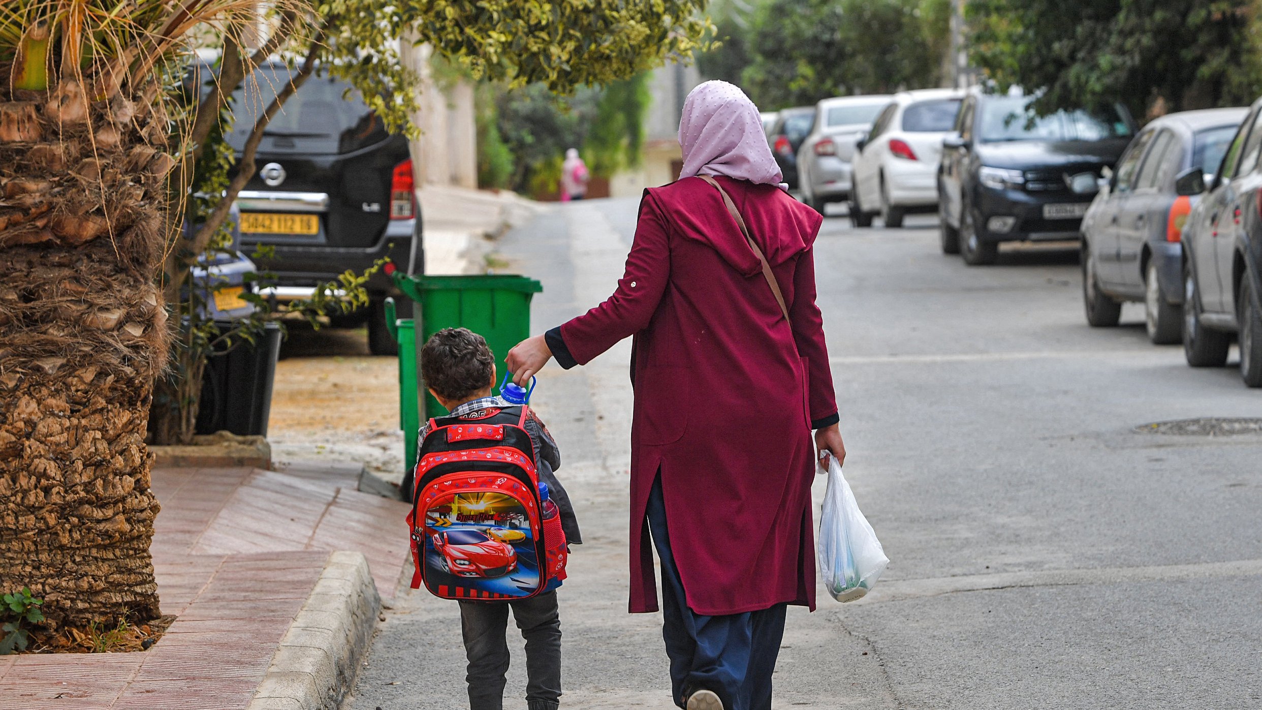 تظهر في الصورة امرأة ترتدي حجابًا وتلبس معطفًا طويلًا، تمشي مع طفل صغير يحمل حقيبة مدرسية على ظهره. يبدو الطفل سعيدًا وهو يمسك بيد والدته. الشارع محاط بالأشجار والمنازل، وهناك سيارات متوقفة على الجانبين. الأجواء تبدو هادئة وتعكس لحظة يومية من حياة الأسرة.
