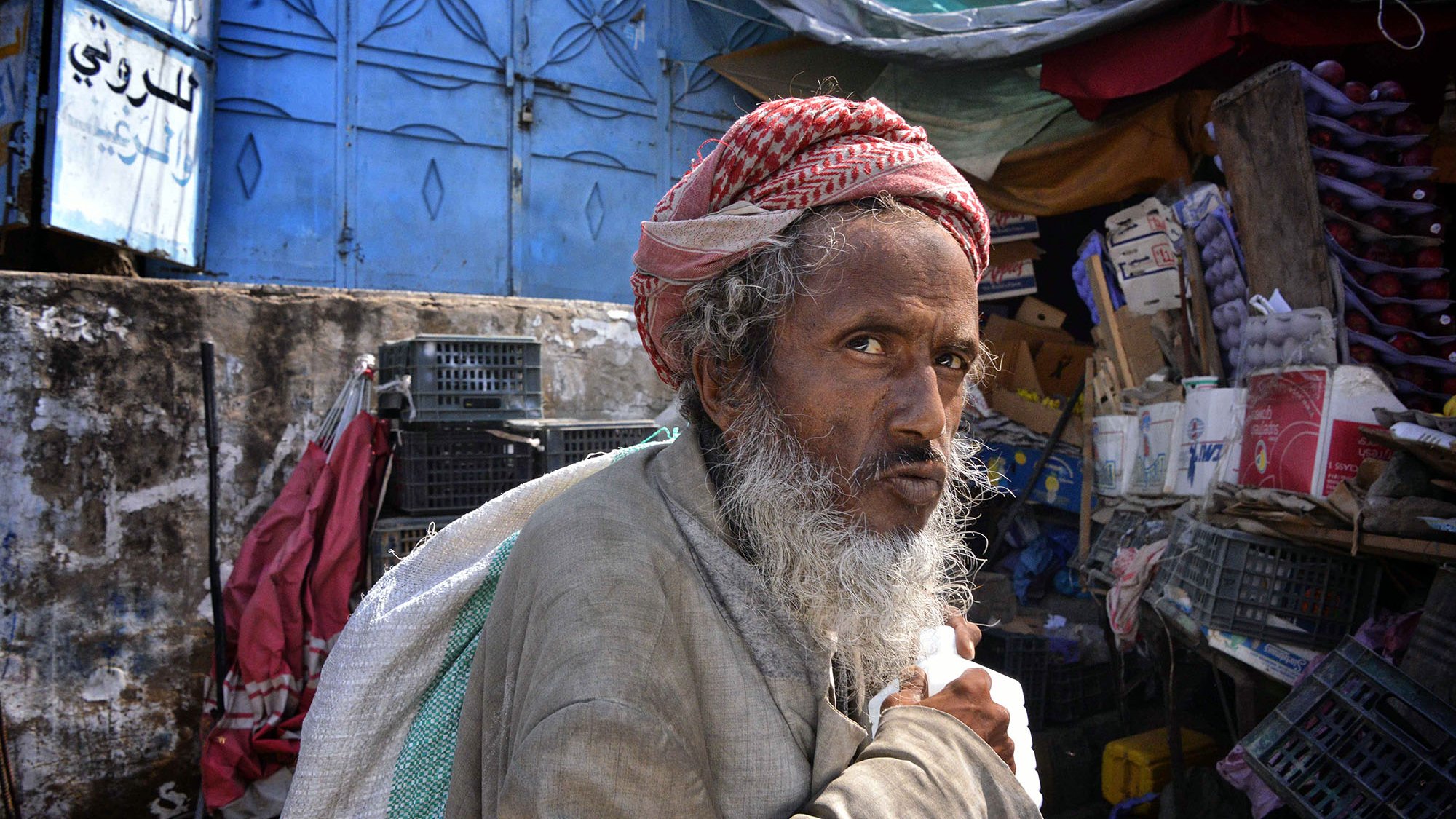 الصورة تُظهر رجلًا مسنًا، يبدو من ملامحه أنه يحمل تجاعيد تعبر عن تجارب الحياة. لديه لحية بيضاء وشعر تحت الغطاء الذي يضعه على رأسه. يرتدي ملابس بسيطة قديمة. تعكس الخلفية بساطتها وفوضاها، حيث توجد حاويات وخزائن وألوان متداخلة. تعبير وجهه يحمل مزيجًا من الحزن والتأمل، وكأنه يعيش في عالم خاص به.