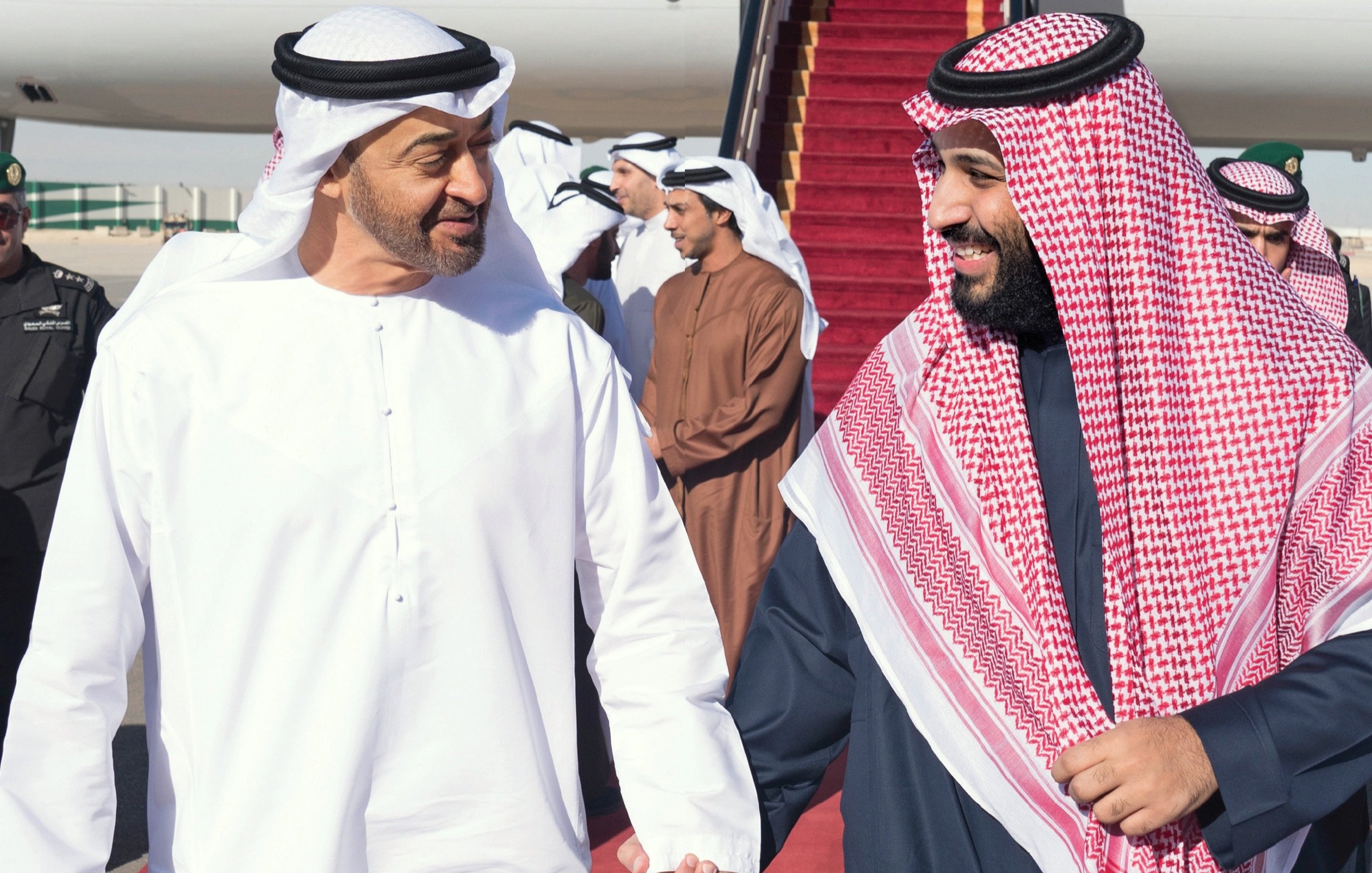 L'image montre deux hommes vêtus de vêtements traditionnels arabes, marchant côte à côte sur un tapis rouge. Ils semblent échanger un moment amical, souriant l'un à l'autre. En arrière-plan, plusieurs autres personnes sont présentes, ainsi qu'un avion stationné. L'atmosphère semble chaleureuse et détendue.