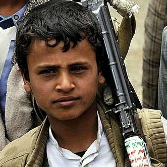 L'image montre un jeune garçon, probablement dans un contexte difficile. Il porte des vêtements simples et a un fusil en bandoulière. Son regard est sérieux, et il semble faire partie d'un groupe plus large, qui est à l'arrière-plan. Les vêtements des autres personnes indiquent un environnement rural ou de conflit. L'atmosphère générale de l'image témoigne d'un contexte de tension.