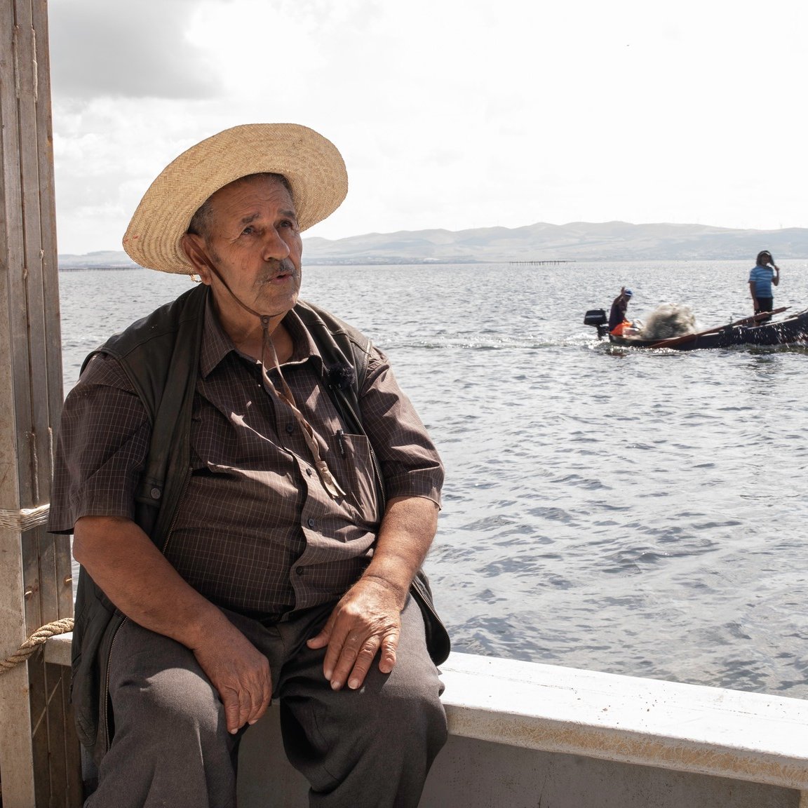 L'image montre un homme assis sur le bord d'un bateau, portant un chapeau de paille large. Il a l'air pensif et regarde vers le large. En arrière-plan, on aperçoit un autre bateau avec des personnes qui semblent s'affairer à la pêche. L'environnement est calme, avec des eaux calmes et un ciel légèrement nuageux, suggérant une ambiance paisible.