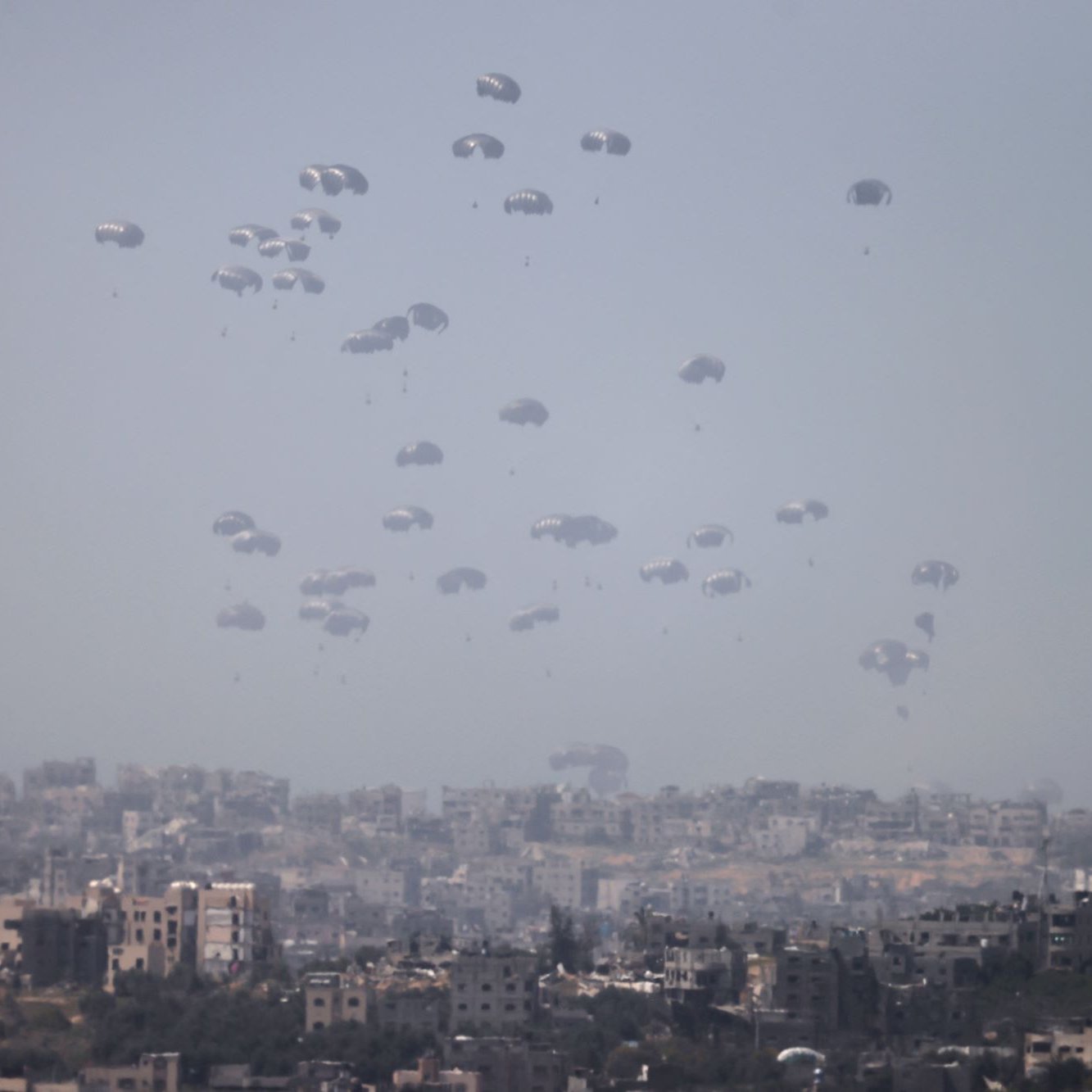 L'image montre un paysage urbain où de nombreux parachutes descendent lentement vers le sol. On peut voir une ville en arrière-plan, avec des bâtiments disposés de manière variable. L'atmosphère semble brumeuse, ce qui donne un aspect à la fois dramatique et intrigant à la scène. Les parachutes indiquent probablement une opération militaire ou humanitaire, créant un contraste avec l'environnement urbain.