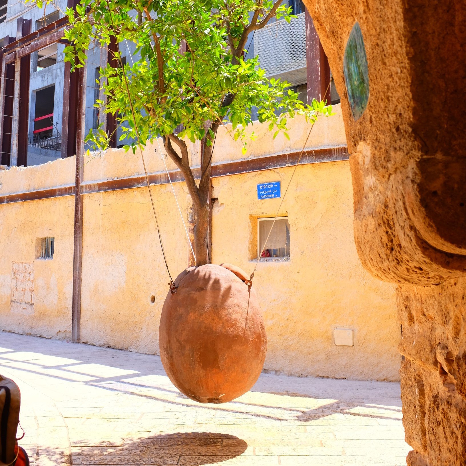 L'immagine mostra un quartiere caratteristico con un muro di colore giallo chiaro. Al centro, si vede un grande vaso di terra cotta che oscilla, sospeso con delle corde. Il vaso sembra appeso a un albero, mentre sullo sfondo si intravede un edificio con strutture in legno e finestre. La scena appare luminosa e vivace, con una sensazione di atmosfera calda e accogliente.