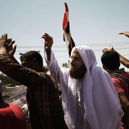L'image montre une scène de rassemblement ou de manifestation. On peut voir un groupe de personnes levant les bras, probablement en signe de protestation ou de célébration. Parmi eux, un homme se distingue par sa tenue traditionnelle, portant un habit clair et un foulard, tout en brandissant un drapeau. L'ambiance semble être celle d'une grande mobilisation, avec des expressions de détermination ou d'engagement sur les visages des participants. Des bâtiments sont visibles en arrière-plan, suggérant que l'événement se déroule en milieu urbain.