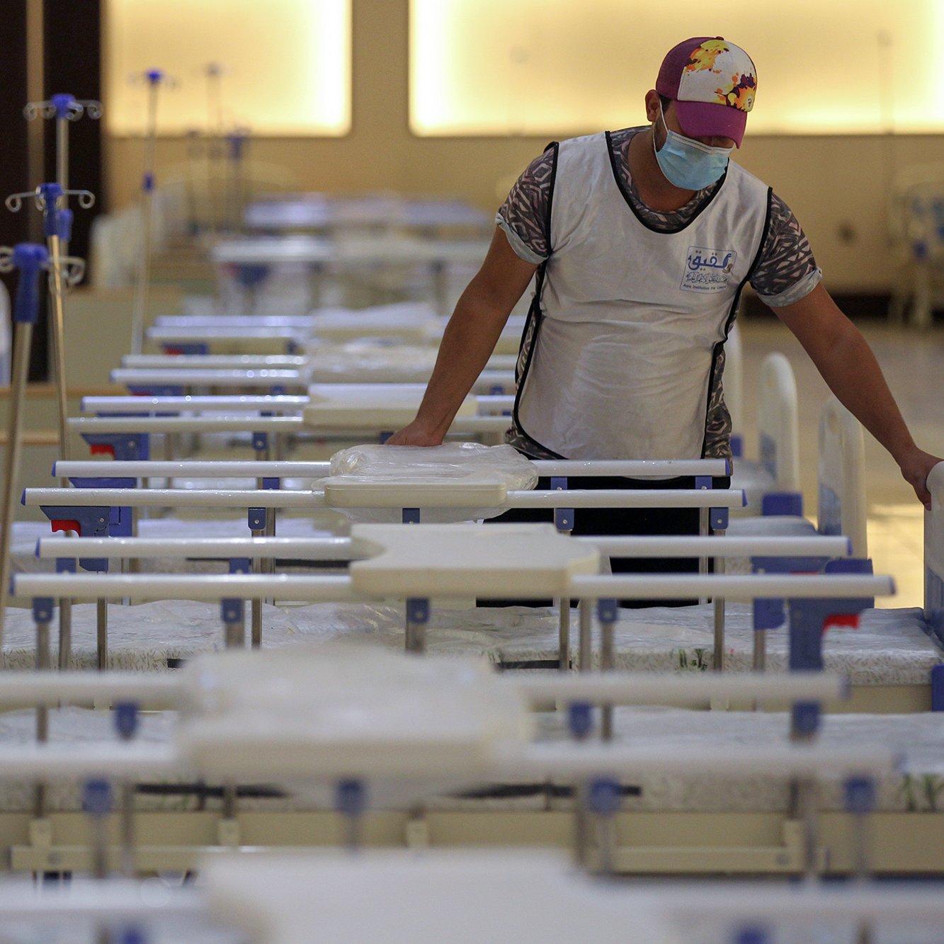 The image shows a person in a mask and a cap standing in a room filled with hospital beds. The beds are arranged in neat rows, and the area appears organized and prepared, possibly for medical purposes. The individual is interacting with the beds, which may indicate they are setting up or maintaining the space. The lighting in the room is warm and soft, contributing to a calm atmosphere.
