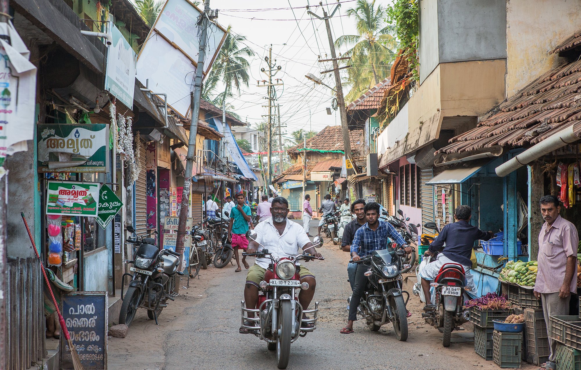 The image depicts a bustling street scene, likely in a South Asian locale. There are narrow, lined pathways with a mixture of residential and commercial buildings, featuring traditional architecture. People can be seen engaging in everyday activities, some on foot and others riding motorcycles. Shops are displaying various goods, and there are signs in a local language. The atmosphere appears lively, with palm trees in the background enhancing the tropical feel of the setting.