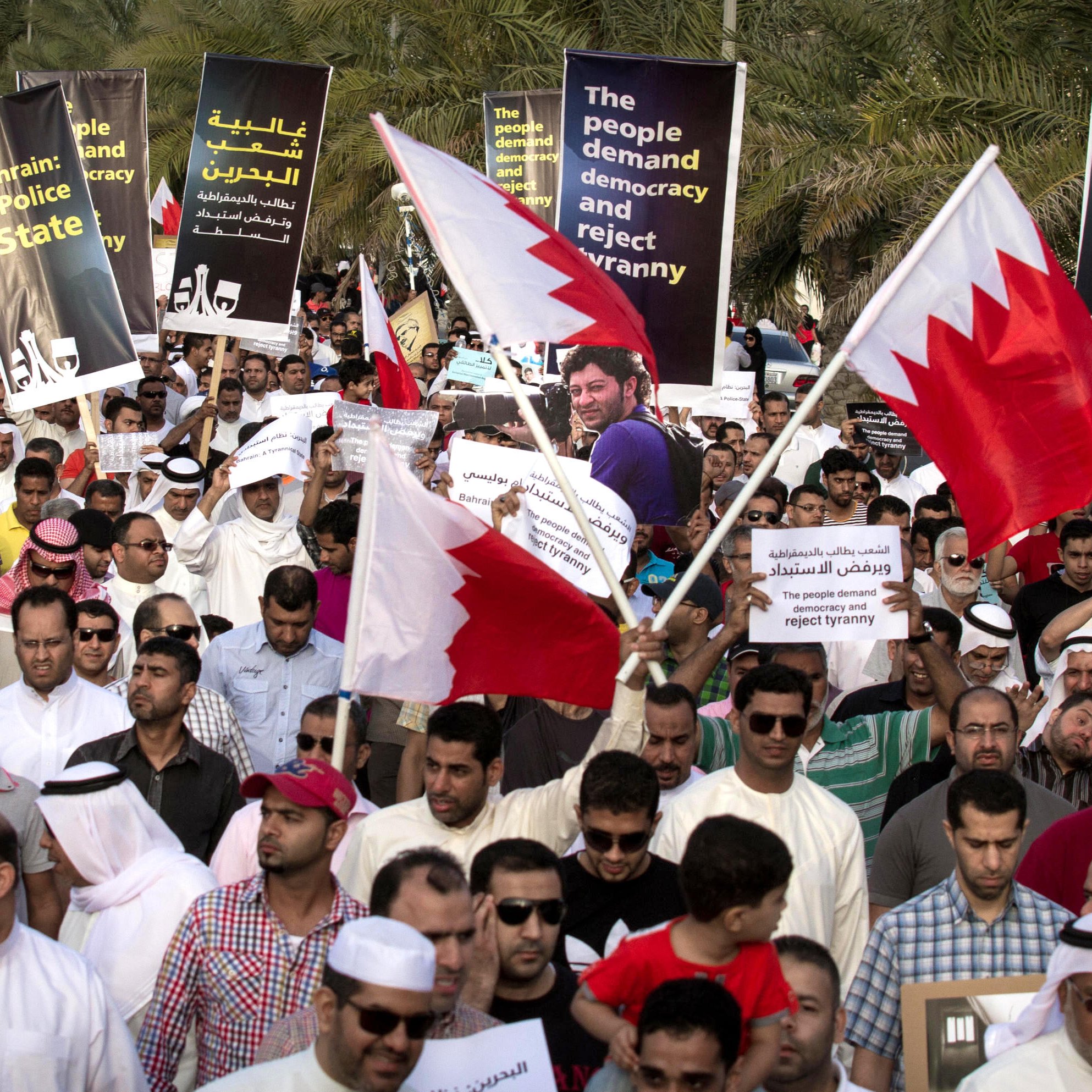L'image montre une grande foule de manifestants rassemblés dans une rue, tenant des pancartes et des drapeaux de Bahreïn. Les manifestants semblent exprimer des revendications politiques, demandant la démocratie et critiquant le régime en place. On peut voir des gens de différentes origines et genres, tous unis dans leur volonté de changement, avec des expressions déterminées sur leurs visages. L'environnement témoigne d'un climat de mobilisation et de protestation collective.