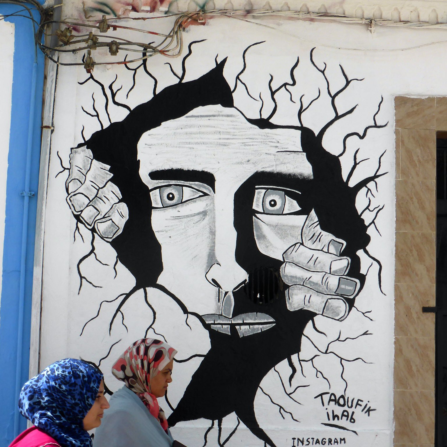 L'image montre une fresque murale en noir et blanc représentant un visage humain au milieu d'une apparente fissure dans le mur. Le visage a des traits marqués et semble exprimer une émotion intense. Des mains émergent des fissures, comme si elles tentaient de sortir. Au premier plan, deux femmes en hijab marchent, apportant une touche de couleur et de vie à la scène. Le tout crée un contraste entre l'art mural et la réalité environnante.