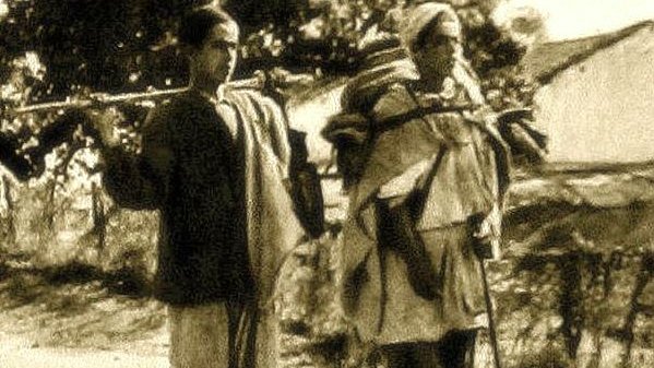 L'image montre deux personnes marchant sur un chemin. L'une d'elles est un jeune garçon, tandis que l'autre est une femme plus âgée. Les deux portent des charges sur leurs épaules, indiquant qu'ils transportent des provisions ou des biens. Le décor semble rural, avec quelques arbres en arrière-plan, et l'atmosphère générale donne un aperçu de la vie quotidienne. Le style de vêtements et le traitement de la photo évoquent une époque passée.