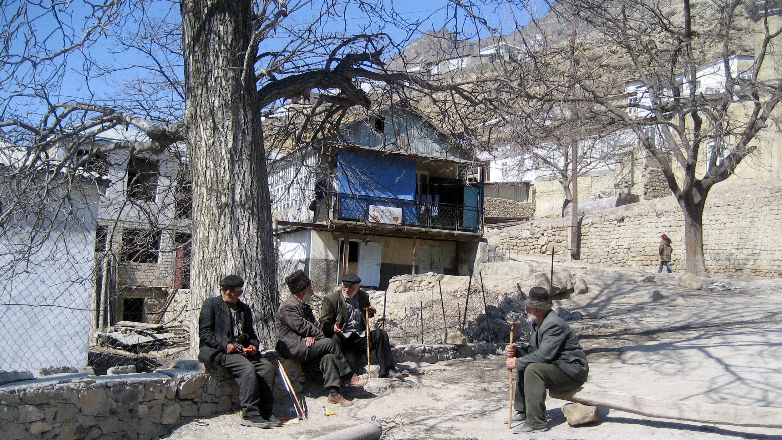 L'image montre un paysage rural avec des maisons en pierre, certaines d'entre elles ayant des balcons. Au premier plan, quatre hommes assis discutent ensemble, habillés de manière traditionnelle. Ils sont entourés d'arbres dénudés, ce qui indique probablement le début du printemps ou l'hiver. L'ambiance semble calme et tranquille, avec un chemin en terre qui serpente entre les maisons.