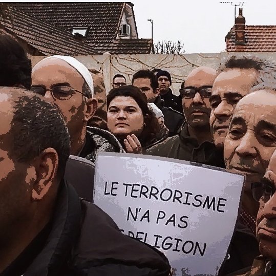 L'image montre un groupe de personnes rassemblées lors d'une manifestation. Ils semblent préoccupés et écoutent attentivement, avec des expressions sérieuses. Certains tiennent des pancartes, dont une qui porte le message "LE TERRORISME N'A PAS DE RELIGION". L'environnement semble urbain, avec des bâtiments en arrière-plan. L'atmosphère suggère une revendication ou une protestation sur un sujet important.