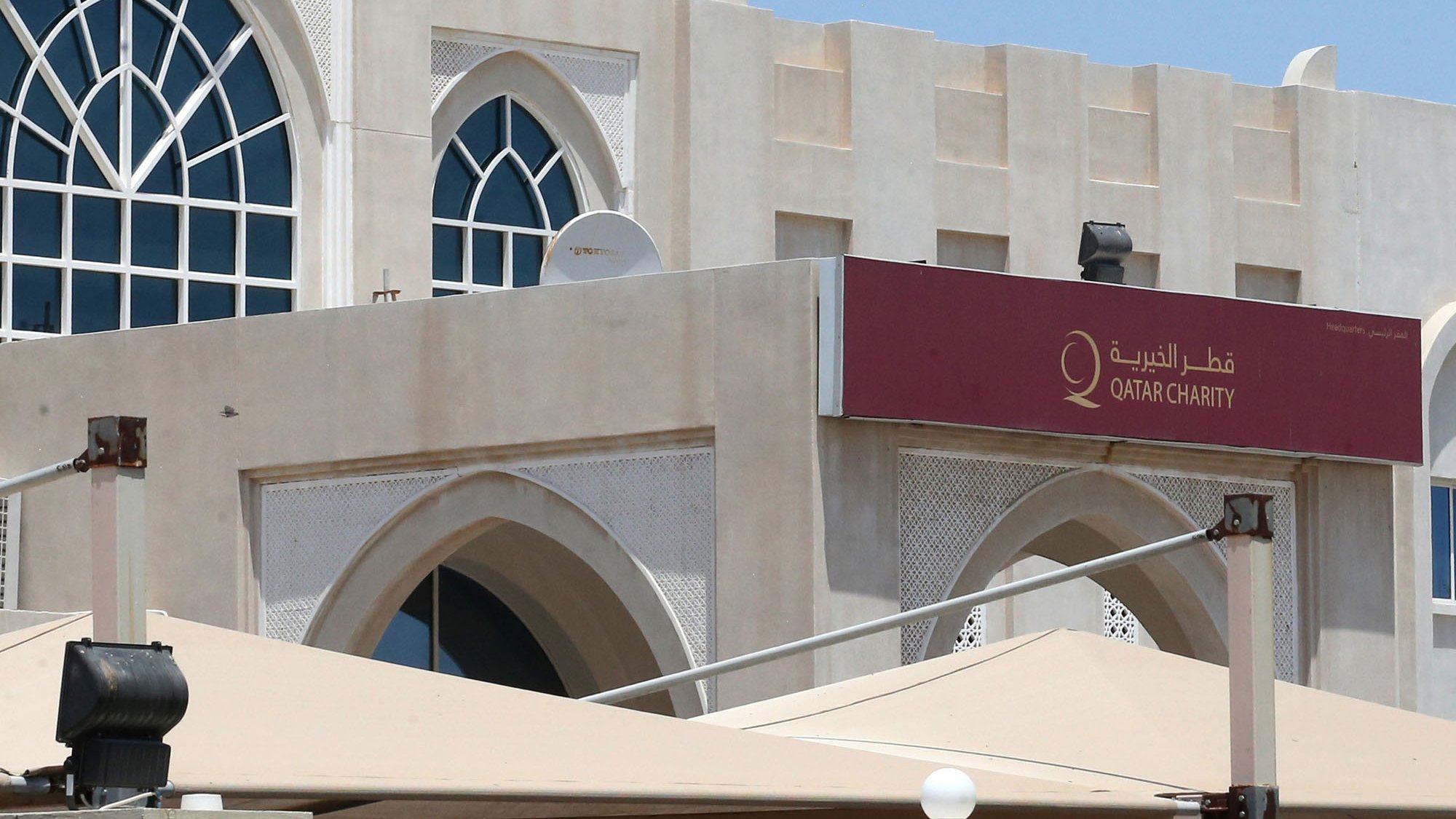 L'image montre un bâtiment de style architectural arabe moderne, avec une grande fenêtre en arc sur le mur. Au-dessus de l'entrée, il y a une enseigne en arabe et en anglais avec le nom "Qatar Charity". Le bâtiment est entouré de parasols et d'une clôture, et on peut voir un drapeau du Qatar flottant au sommet du bâtiment. Le ciel est dégagé et ensoleillé.
