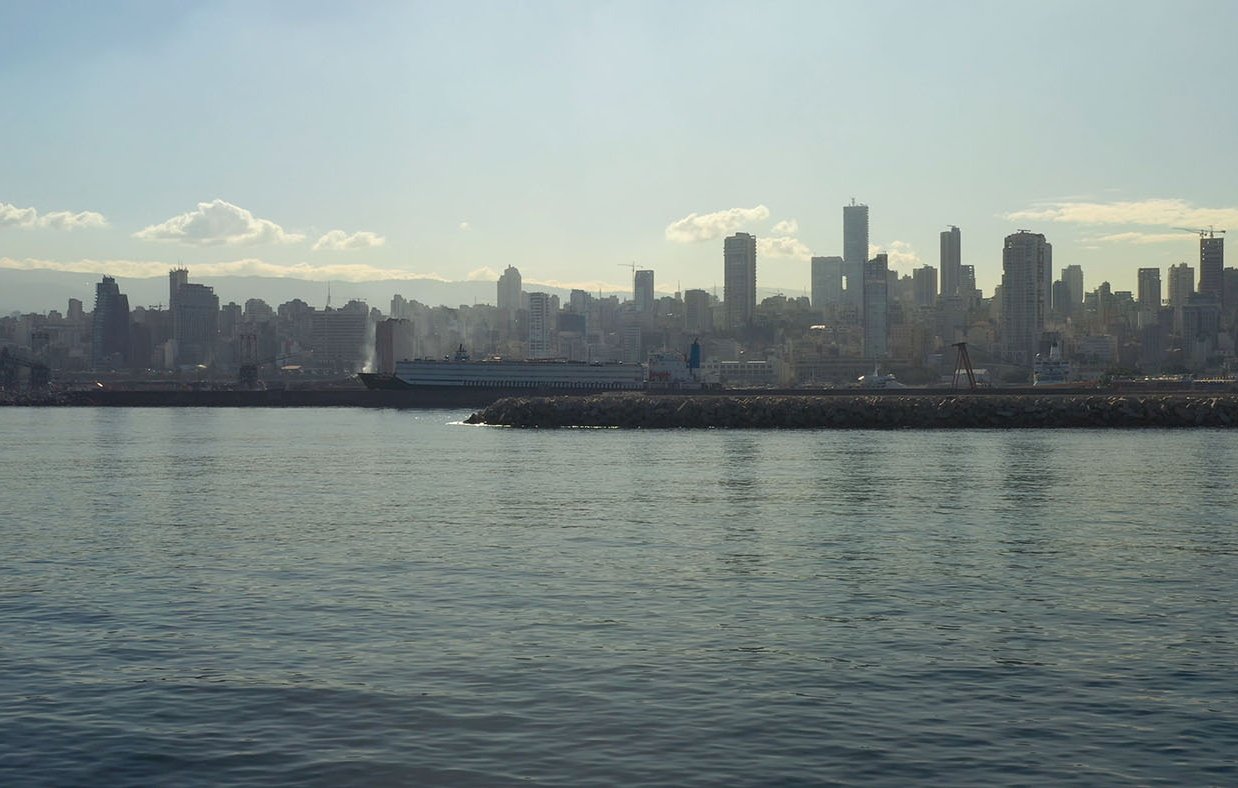 L'image montre un paysage urbain d'une ville moderne en bord de mer. On peut voir des gratte-ciels s'élevant le long de la côte, reflétant la lumière du soleil sur l'eau calme de la mer. Au premier plan, il y a un port ou une jetée, et l'arrière-plan est rempli de bâtiments qui se dressent majestueusement. Le ciel est légèrement nuageux, ce qui crée une ambiance douce et mélancolique. L'ensemble donne une impression de tranquillité et de modernité.