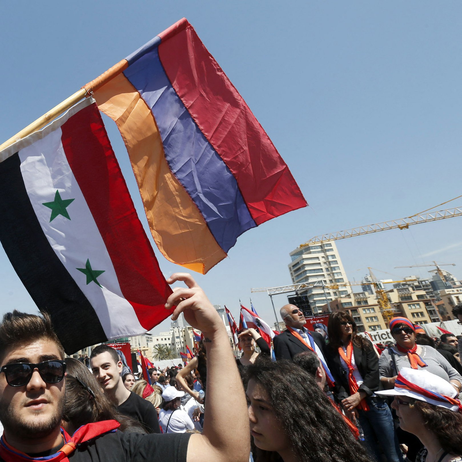 L'image montre un groupe de personnes participant à une manifestation ou un rassemblement. Un homme au premier plan tient fièrement trois drapeaux : le drapeau syrien, un drapeau avec trois étoiles vertes sur fond noir et rouge, ainsi qu'un drapeau arménien. L'ambiance semble festive et engagée, avec de nombreuses personnes en arrière-plan, probablement partageant des sentiments communs ou soutenant une cause. Le ciel est clair, ce qui ajoute à l'énergie de l'événement.