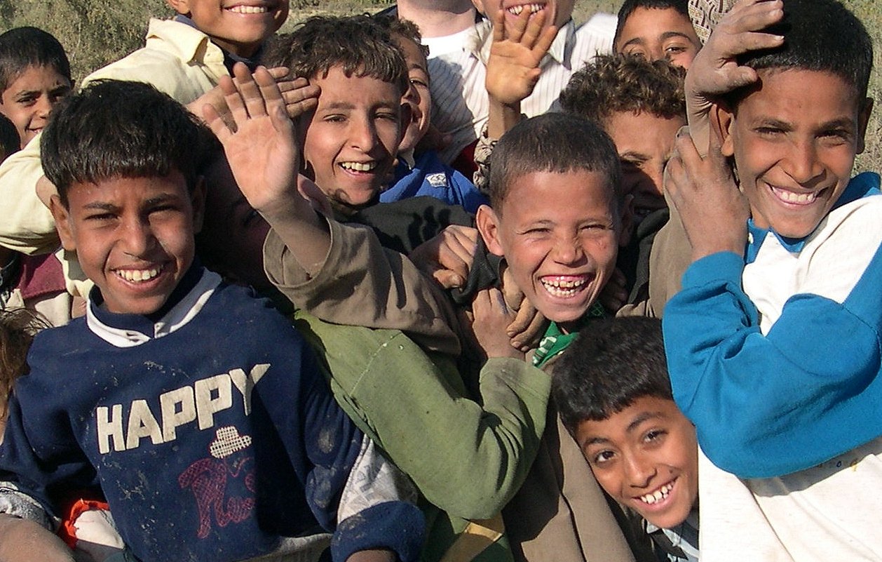 L'image montre un groupe d'enfants qui sourient joyeusement. Ils semblent apprécier le moment, avec des expressions de joie et beaucoup d'énergie. Les enfants sont vêtus de vêtements variés et certains lèvent la main ou se penchent vers l'avant, montrant un sentiment d'excitation et d'unité. L'arrière-plan est flou, mettant en avant leurs visages animés et heureux.