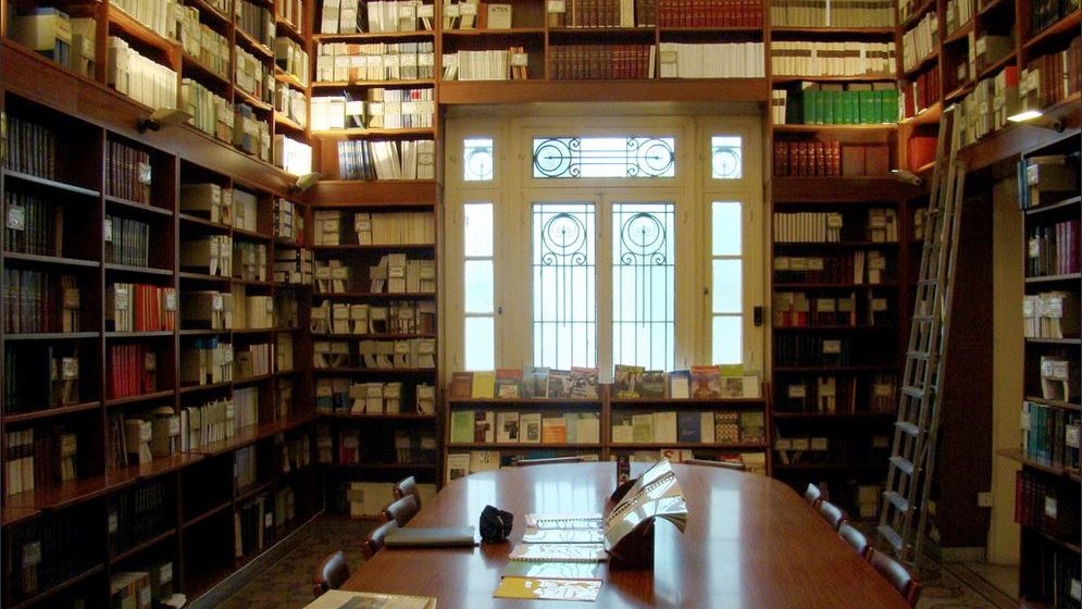 L'image montre une bibliothèque élégante et bien organisée. Les étagères sont remplies de livres sur plusieurs niveaux, allant du sol au plafond. Au centre, il y a une grande table en bois entourée de chaises, créant un espace propice à la lecture ou à l'étude. La lumière naturelle pénètre par de grandes fenêtres avec des motifs décoratifs, apportant une ambiance chaleureuse et accueillante. L'ensemble de l'endroit évoque un sentiment de calme et de sérénité, idéal pour les passionnés de lecture.
