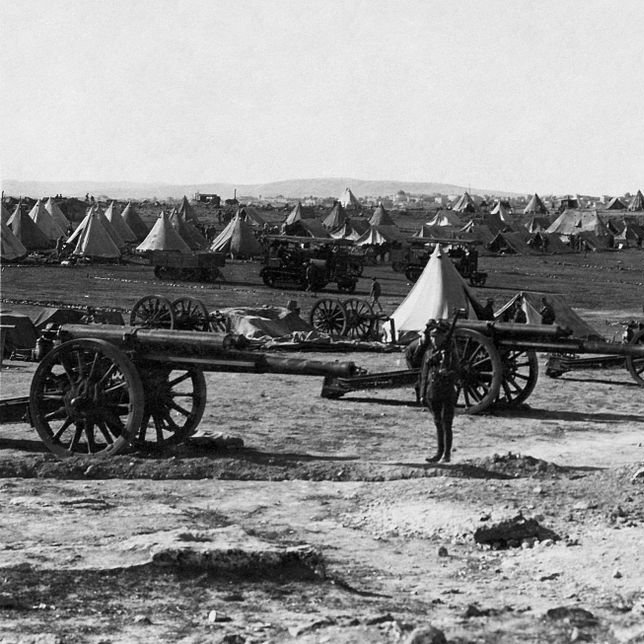 L'image montre un camp militaire avec plusieurs canons en premier plan. À l'arrière-plan, on distingue des tentes disposées de manière ordonnée sur un terrain dégagé. Le paysage semble vaste, avec des collines ou des montagnes au loin. L'atmosphère est historique, évoquant une période de guerre ou de mobilisation des troupes. Un soldat ou un homme se tient près des canons, donnant une échelle à la scène.
